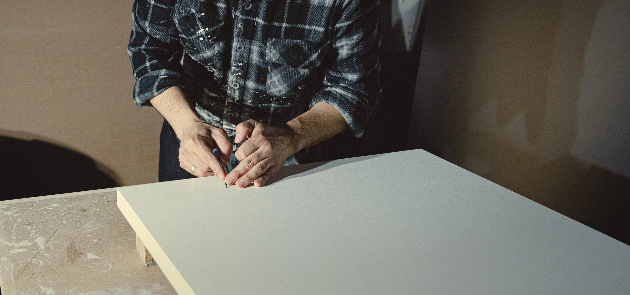 A person wearing a black and gray plaid shirt is using a utility knife to cut a large sheet of white material on a worktable.