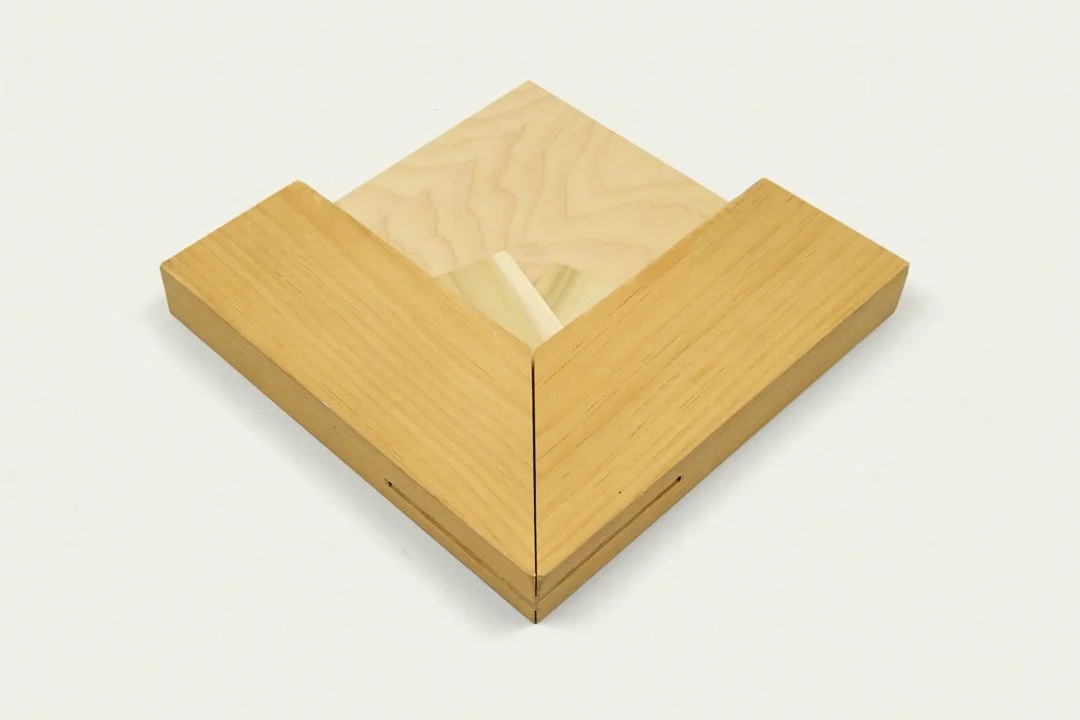 A wooden square corner joint with two visible pieces of light and medium-toned wood on a white background.