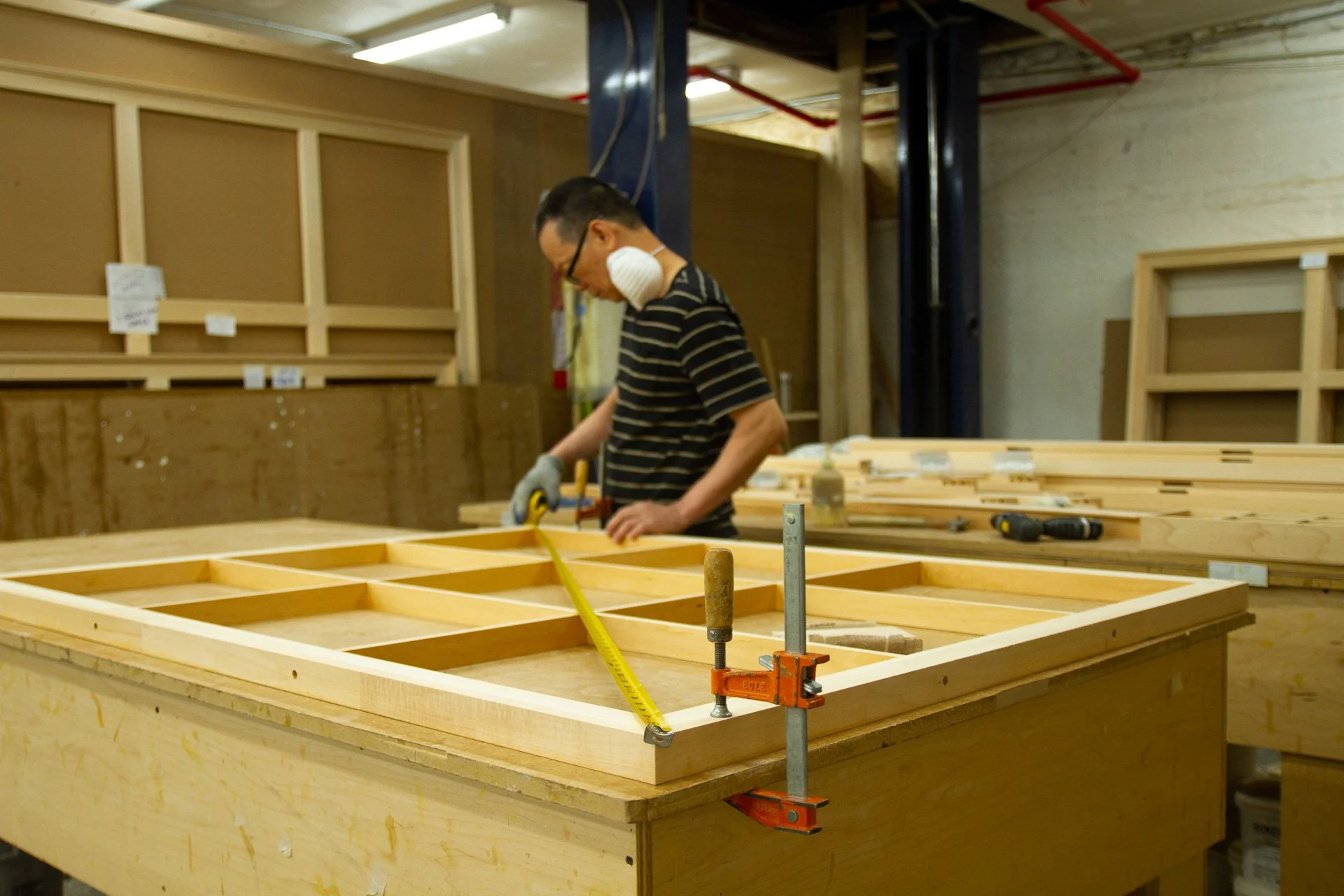 A man wearing safety glasses, a face mask hanging around his neck, and gloves measures a wooden frame with a tape measure at a woodworking shop.
