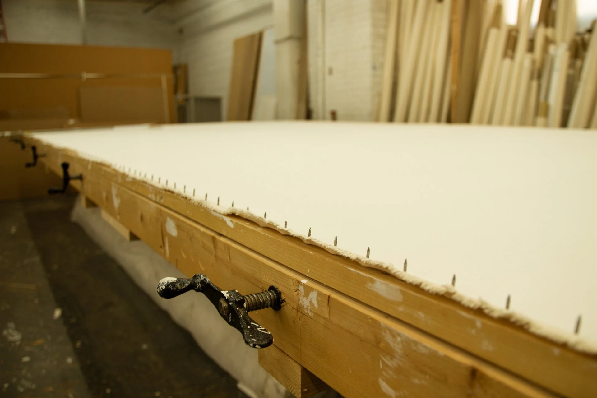 Wooden workbench in woodworking workshop with clamps, surrounded by stacks of canvas and supplies.
