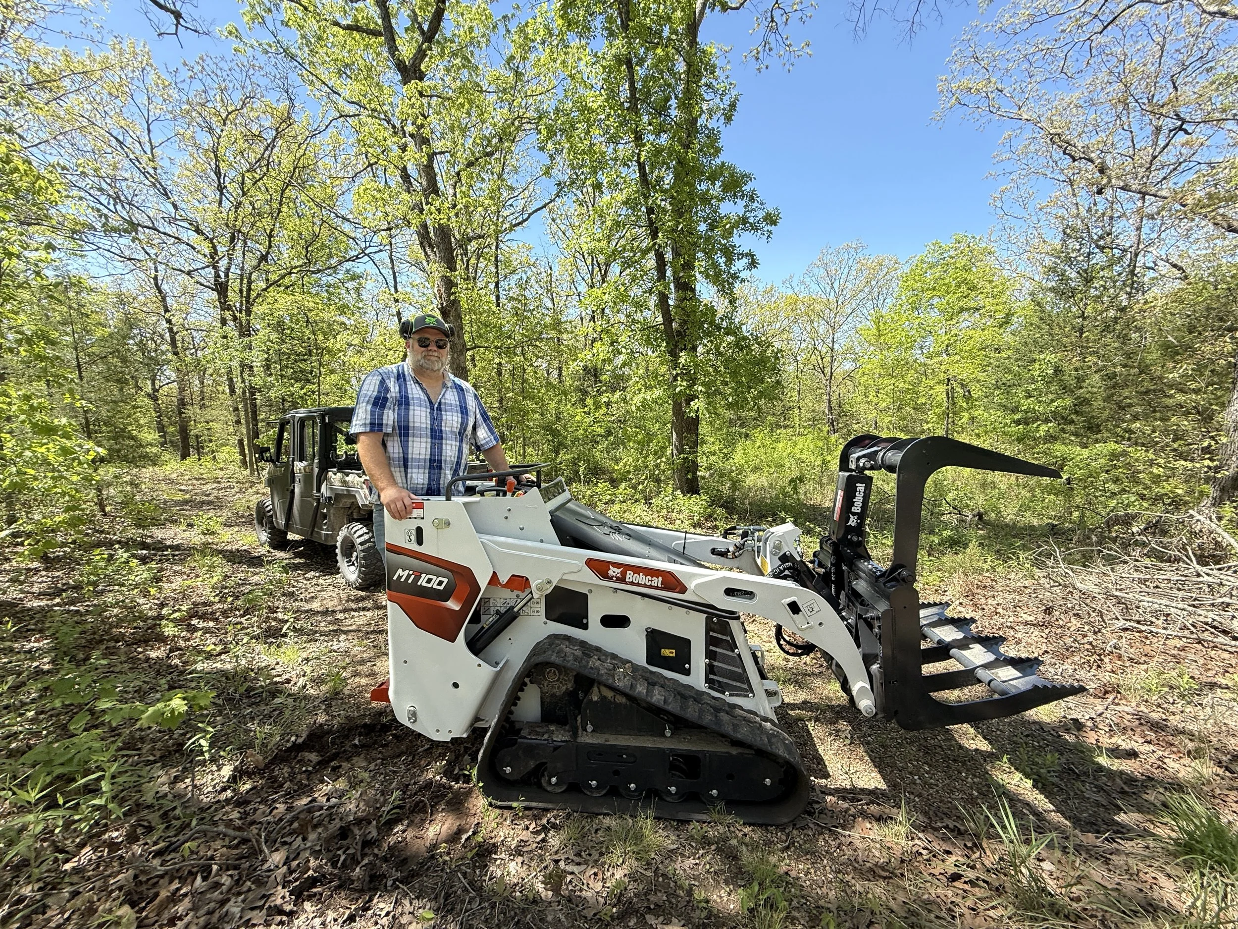 A man in sunglasses and a plaid shirt standing next to a Bobcat machine in a wooded area with trees and blue sky.