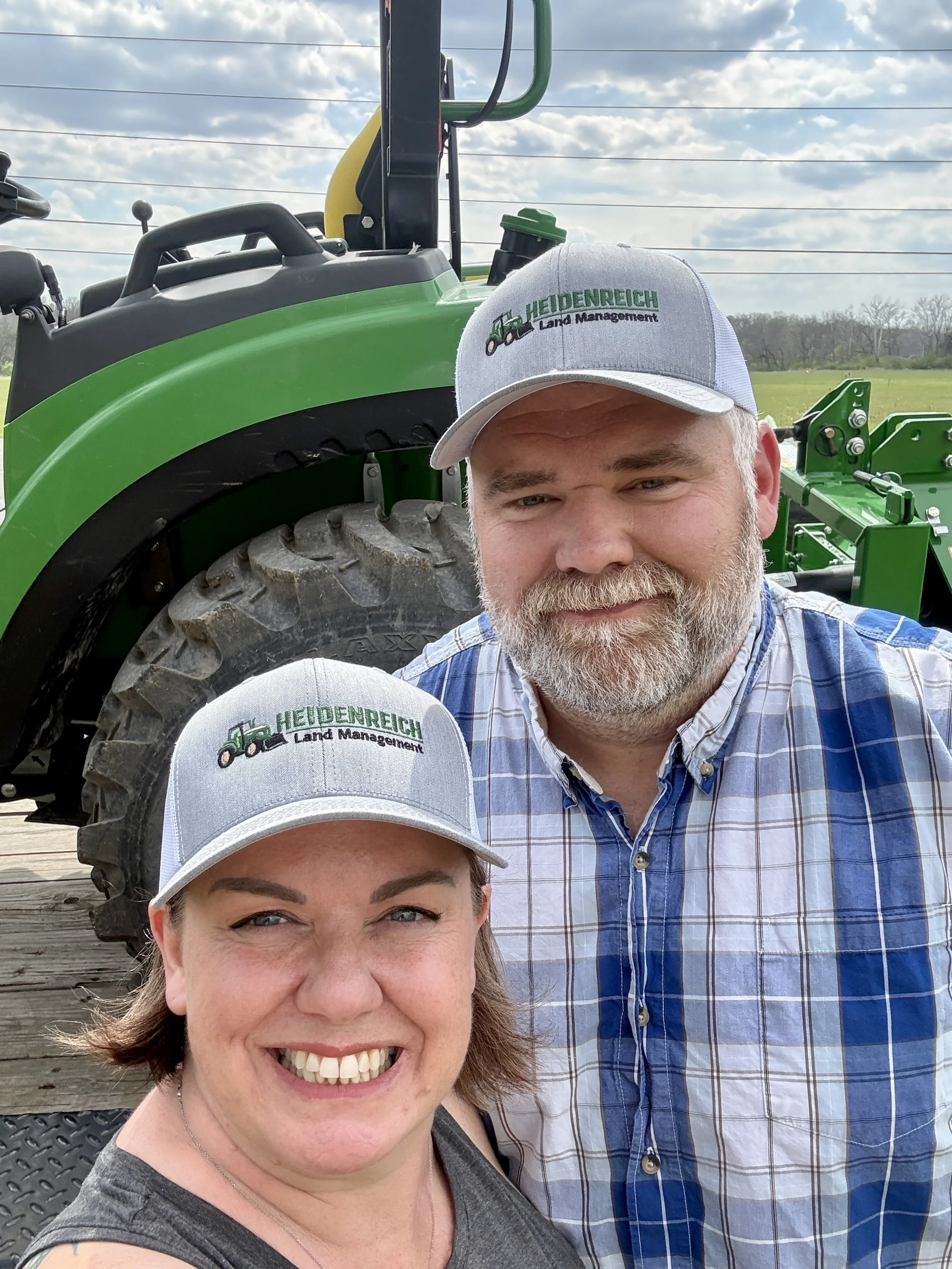 A smiling woman and a man with a beard taking a selfie together outdoors, wearing baseball caps with 'Heidenreich Land Management' logo, with a green tractor and blue sky with clouds in the background.