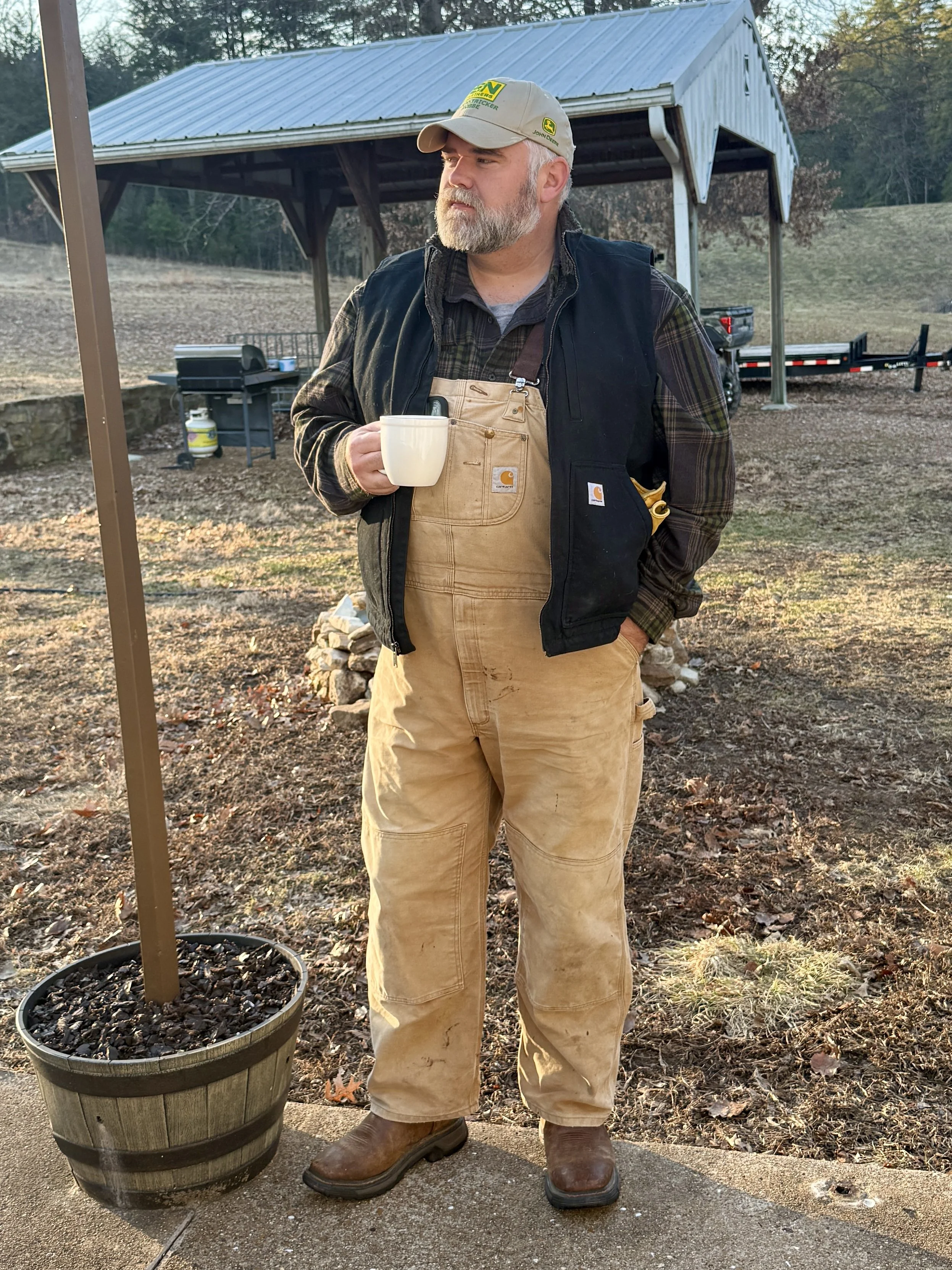 A man standing outdoors holding a white mug. He is wearing a beige cap, a black jacket over a plaid shirt, beige overalls, and brown boots. There is a metal shed in the background with trees and a trailer behind him.