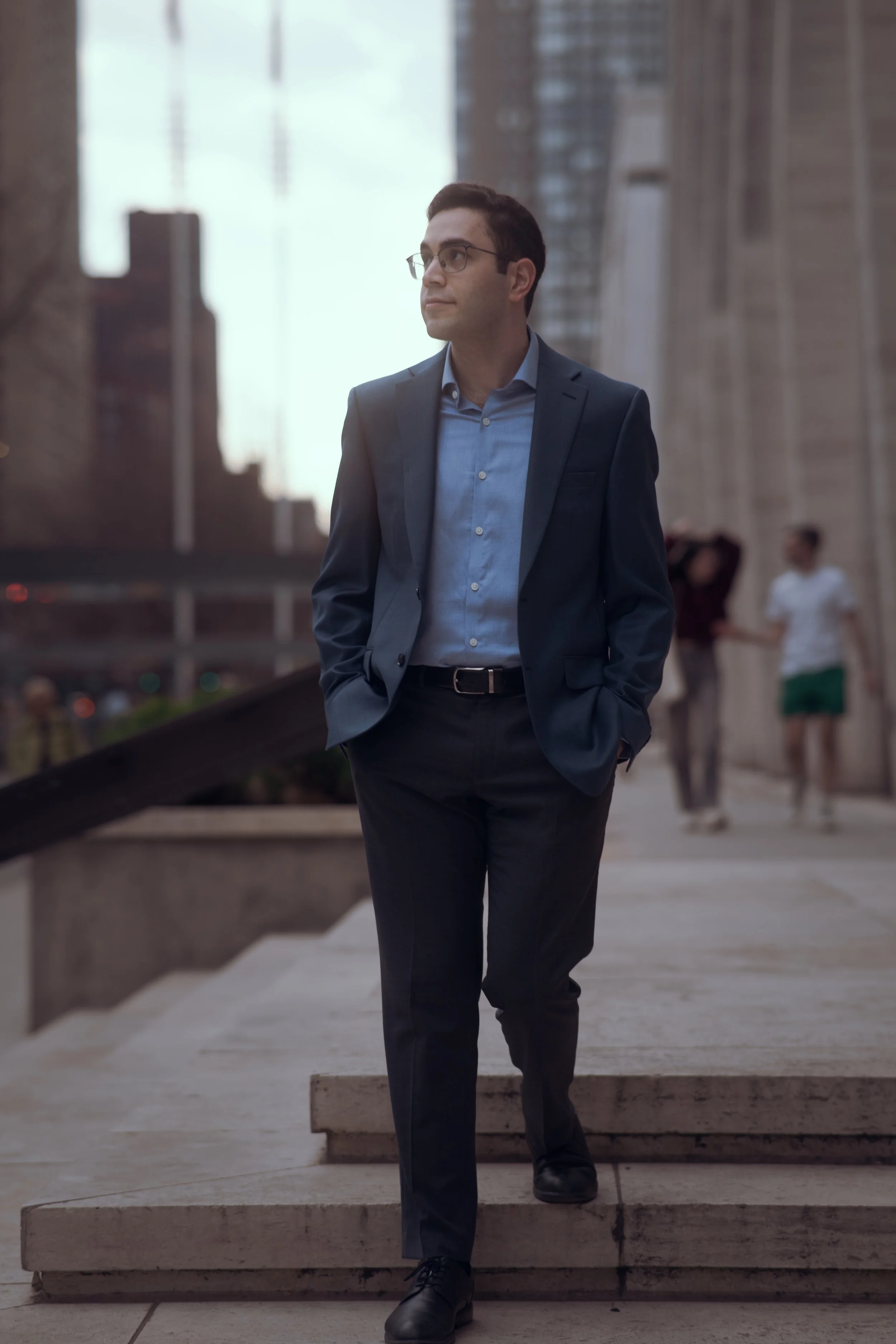 A man in a navy blue suit and glasses walking down stairs outdoors in an urban setting, with tall buildings and blurred pedestrians in the background.
