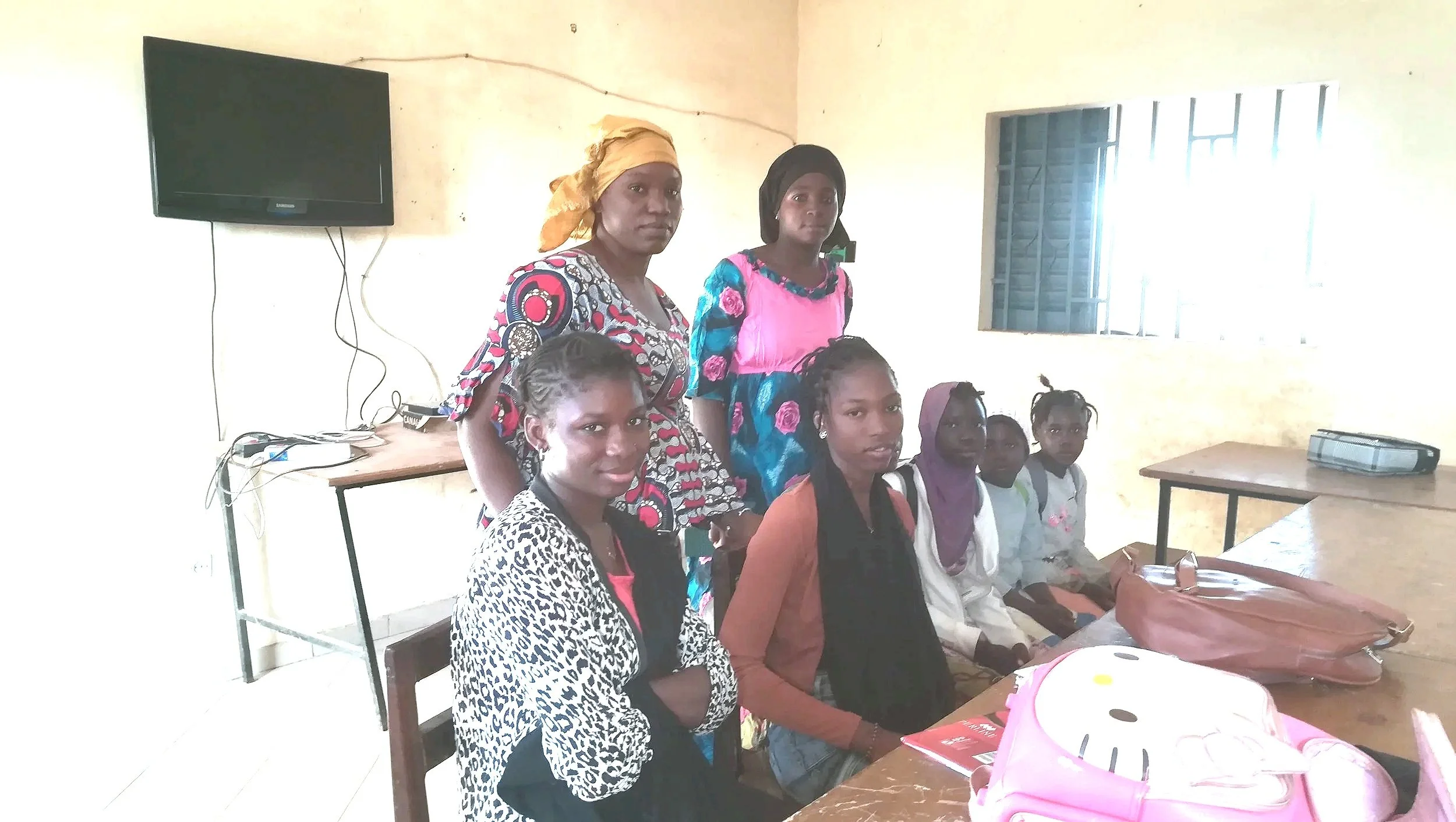A group of women and girls in a classroom in Bamako, Mali, with a television and a table, some sitting and some standing, facing the camera.