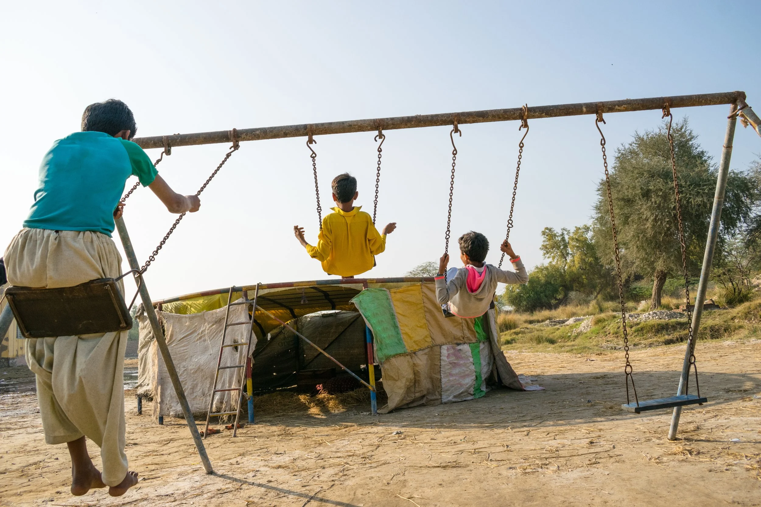 Three children playing on swings in an outdoor area near a makeshift tent, with trees in the background and a clear sky.