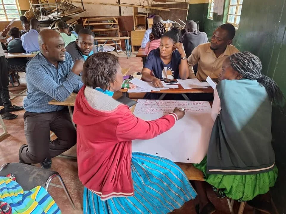 A group of six adults sitting around a table in a classroom in Zambia, engaged in a discussion or workshop, with papers and a large sheet of paper with writing on it.