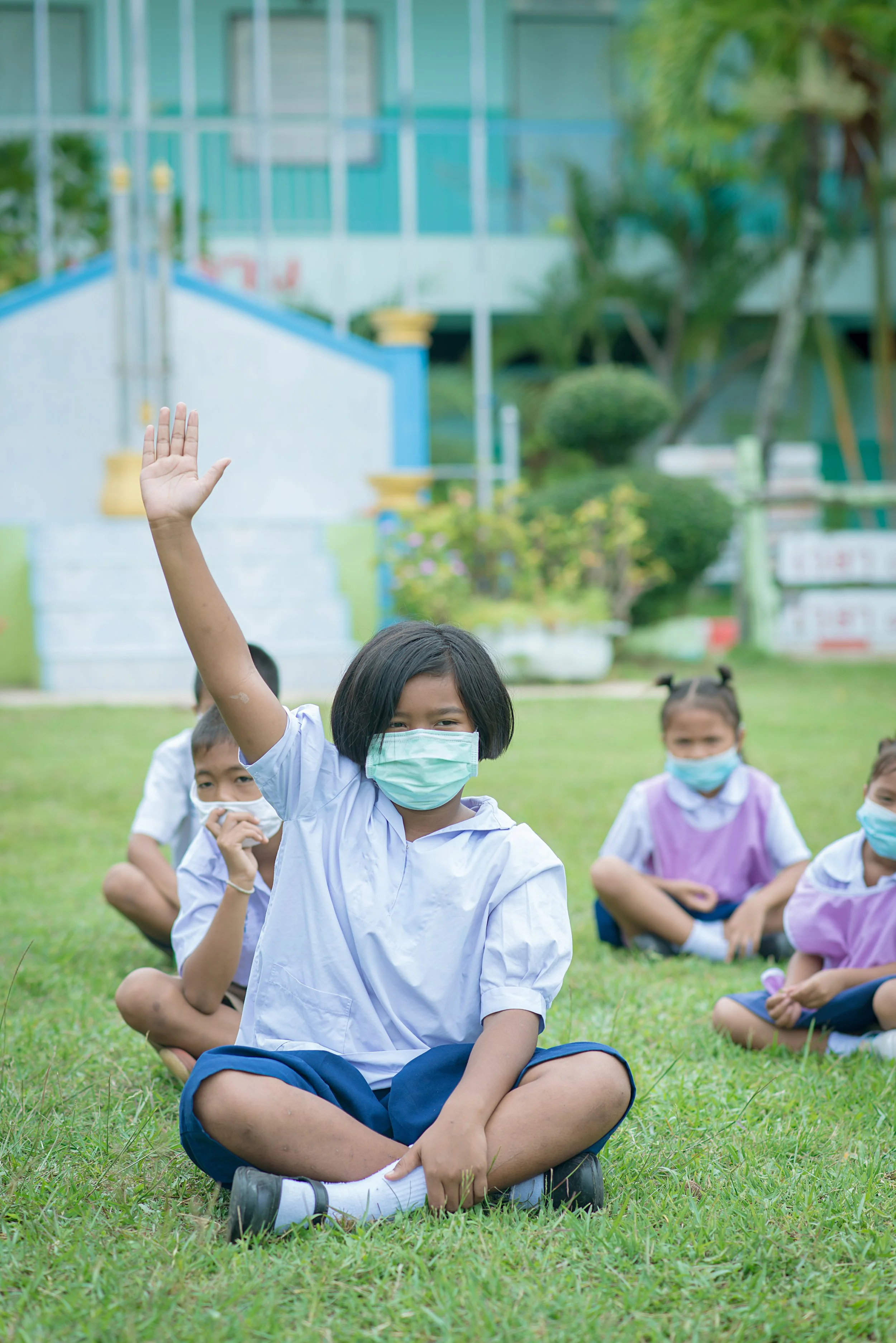 Group of children sitting on grass outdoors, wearing masks, with a girl in the front raising her hand.