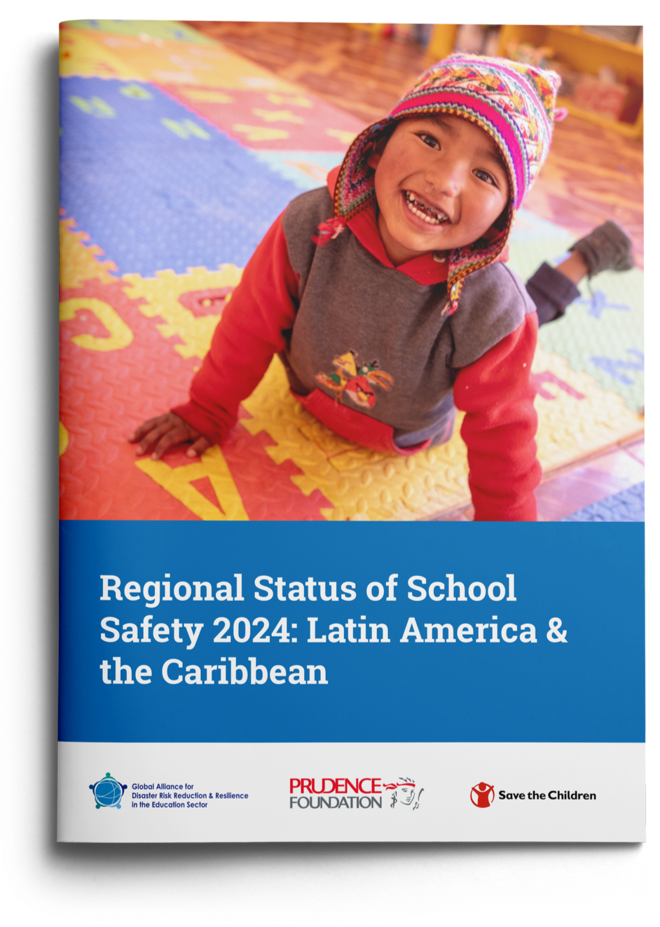 A smiling young child sitting on a colorful foam play mat indoors, wearing a beanie and a gray and red sweatshirt.