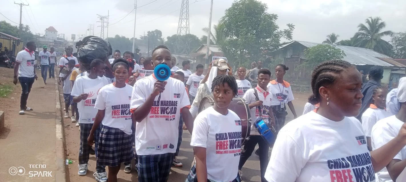 Group of children and adults participating in a march in Liberia for the Child Marriage Free World campaign wearing white t-shirts with campaign messages, walking along a paved road in an urban area.