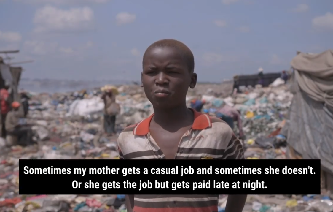 A young boy working as a child labourer standing in a landfill in Kenya with lots of trash and debris around, wearing a striped shirt, under a partly cloudy sky.