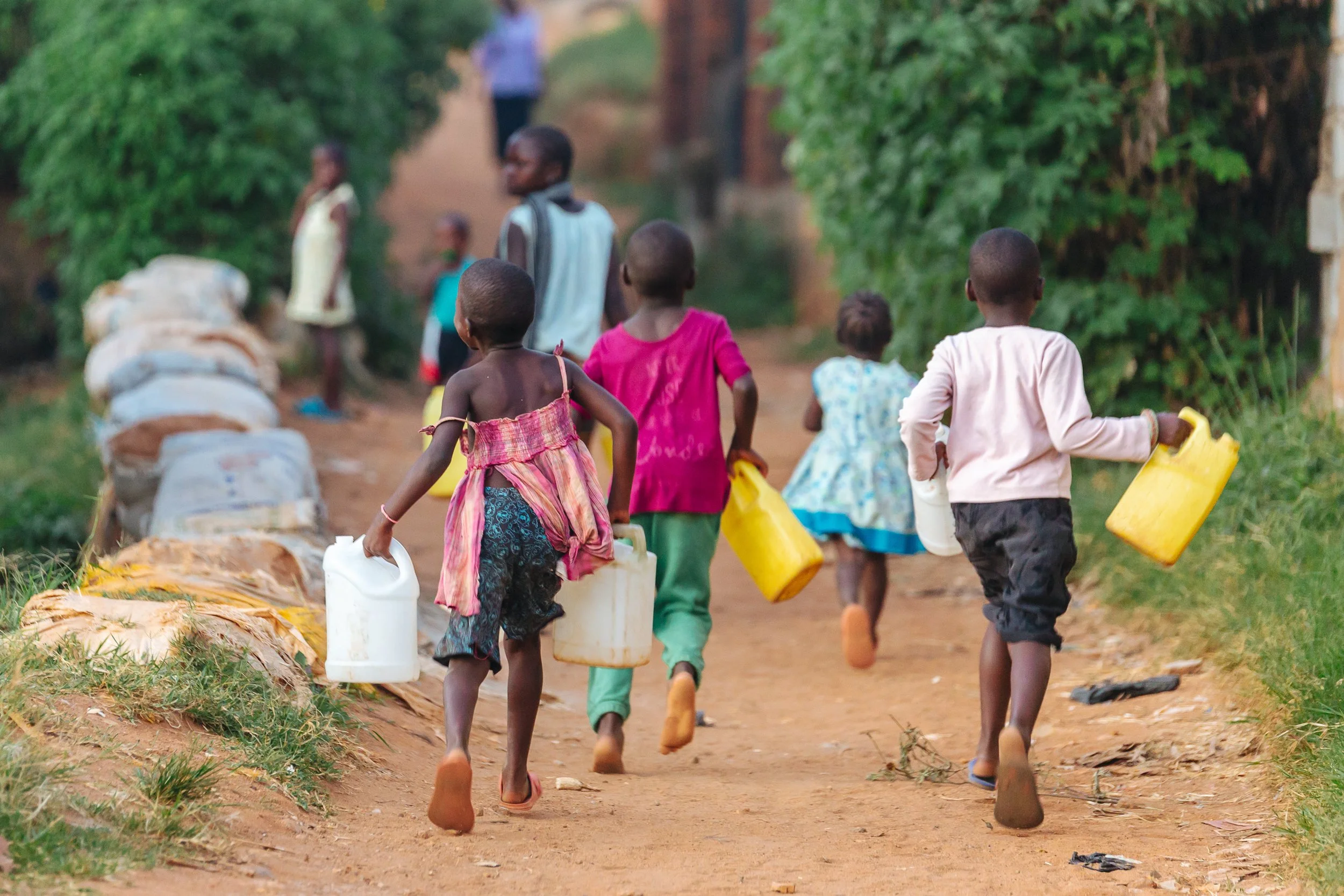 Children carrying water containers walking along a dirt path in a rural area surrounded by greenery.