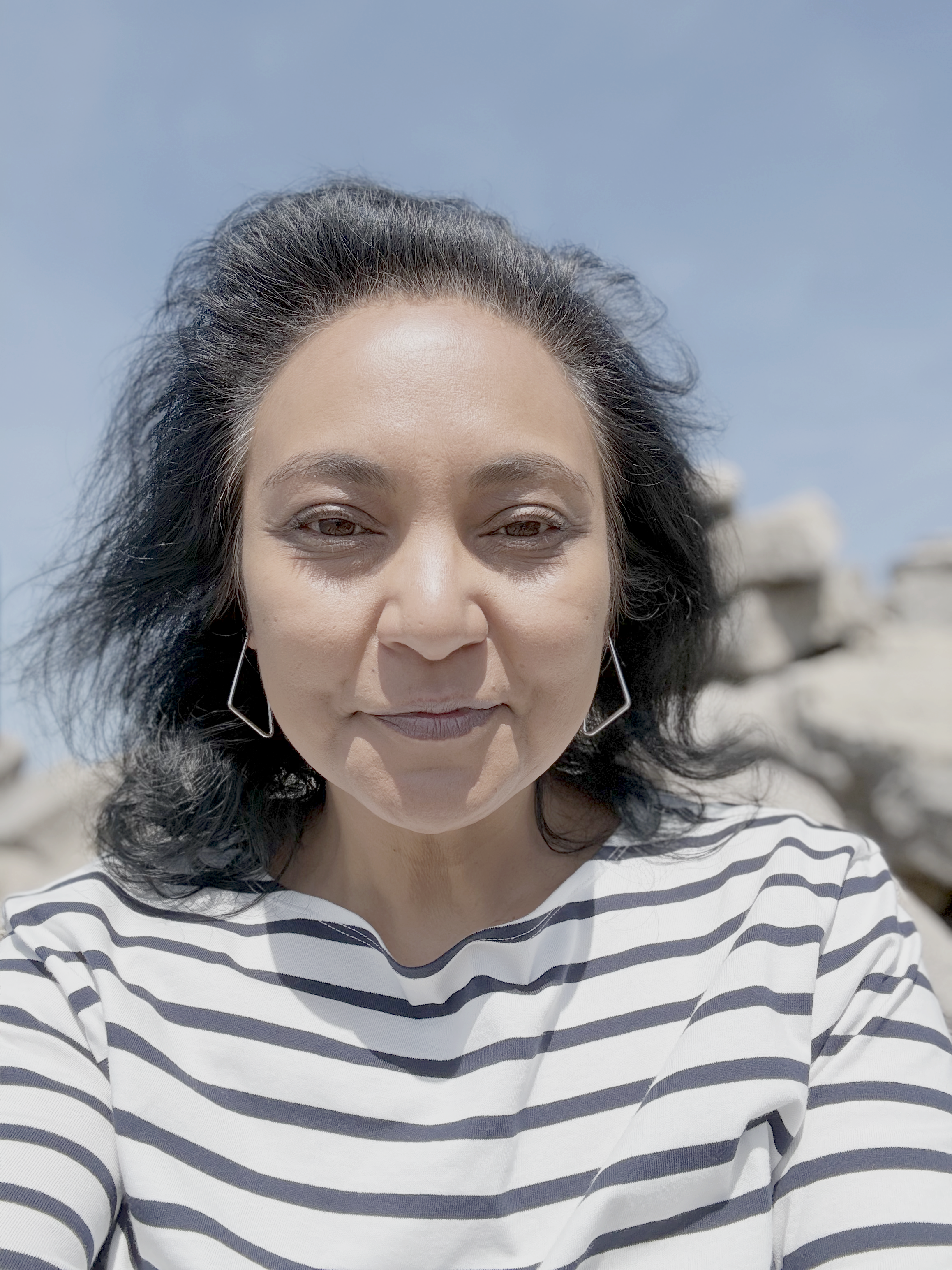 A woman with black hair wearing a striped white and navy shirt outdoors on a sunny day, with rocks and blue sky in the background.