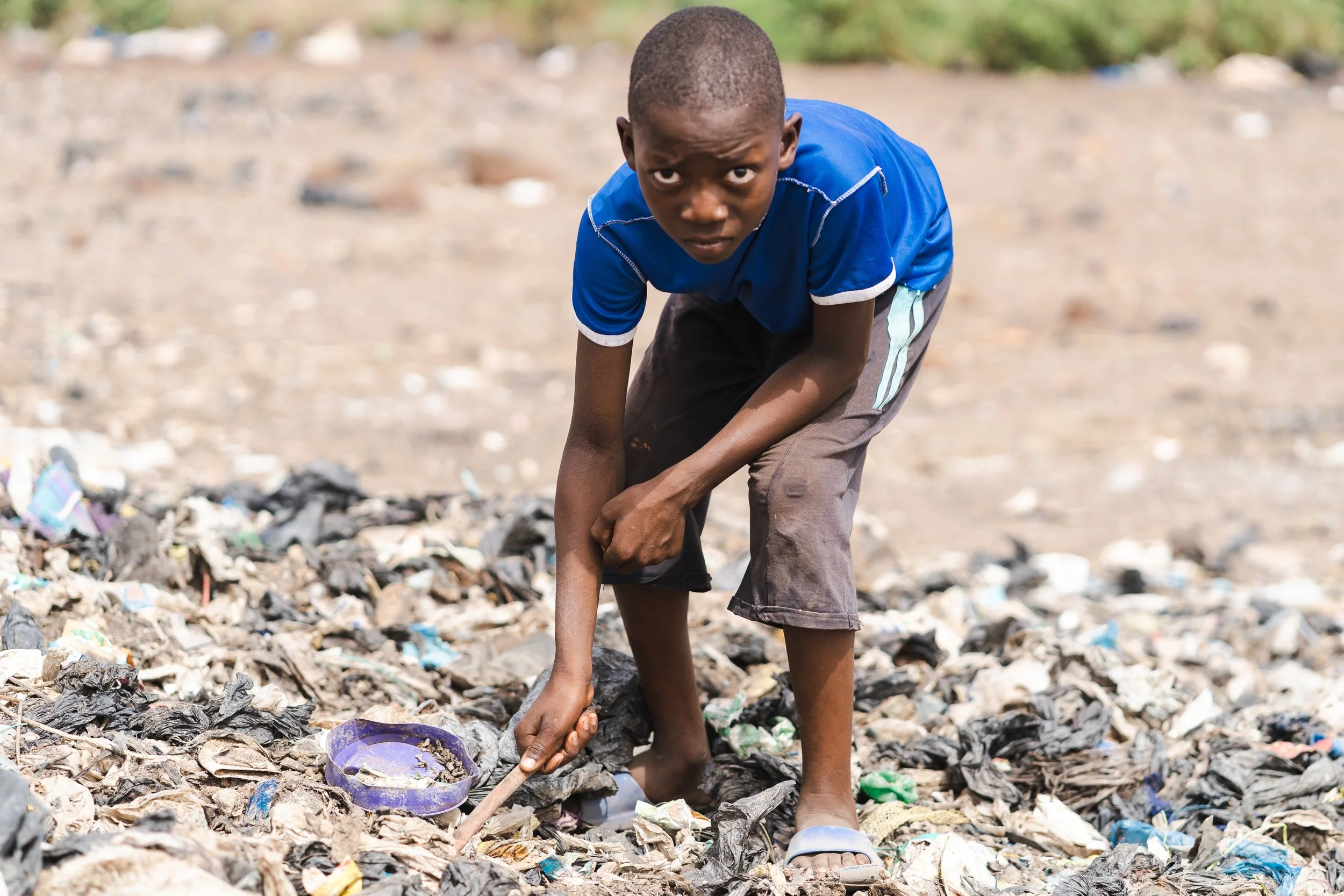 A young boy wearing a blue shirt and shorts is picking trash from a polluted, garbage-filled area with a small spade. The ground is covered with debris and plastic waste.