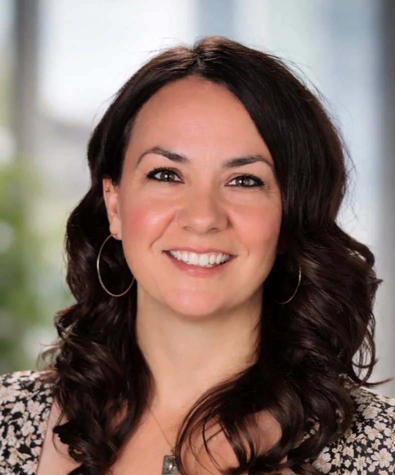 A woman with long dark curly hair smiling, wearing hoop earrings and a patterned top, in a well-lit indoor setting.