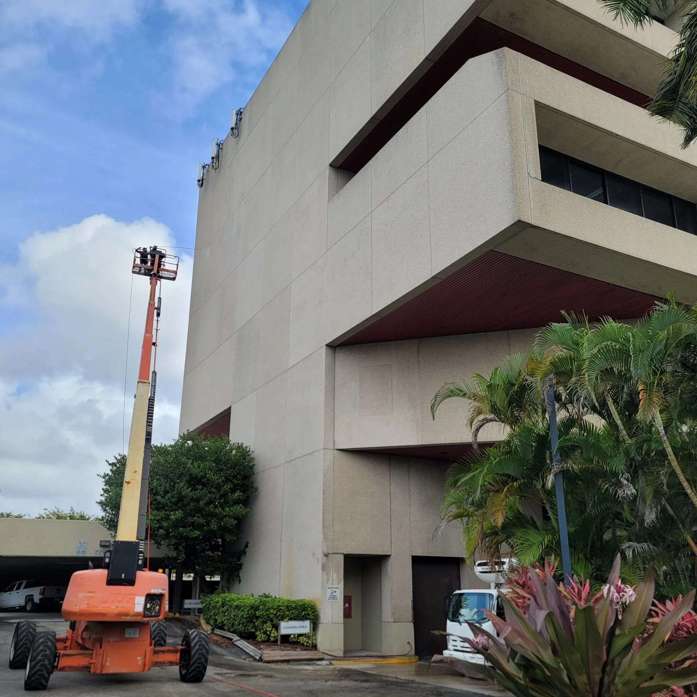 A beige painted multi-story Miami building with brown accents, surrounded by green plants and palm trees. Painting equipment is present for performing paint services, including a lift and some parked vehicles, under a partly cloudy sky.