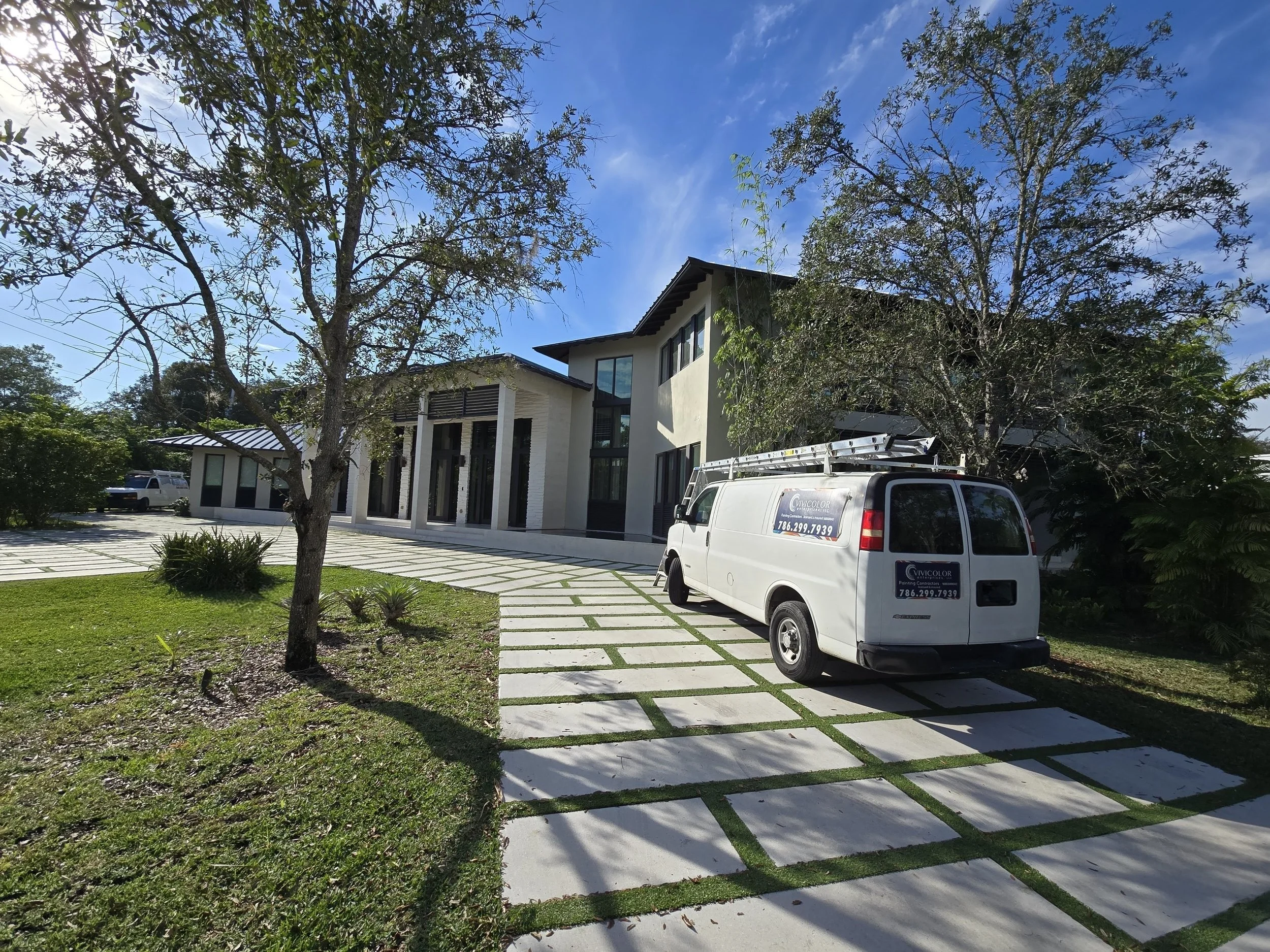 Modern painted Miami house in with a white exterior paint, large windows, and a landscaped front yard with trees and a driveway with pavers. A white paint service van is parked outside.