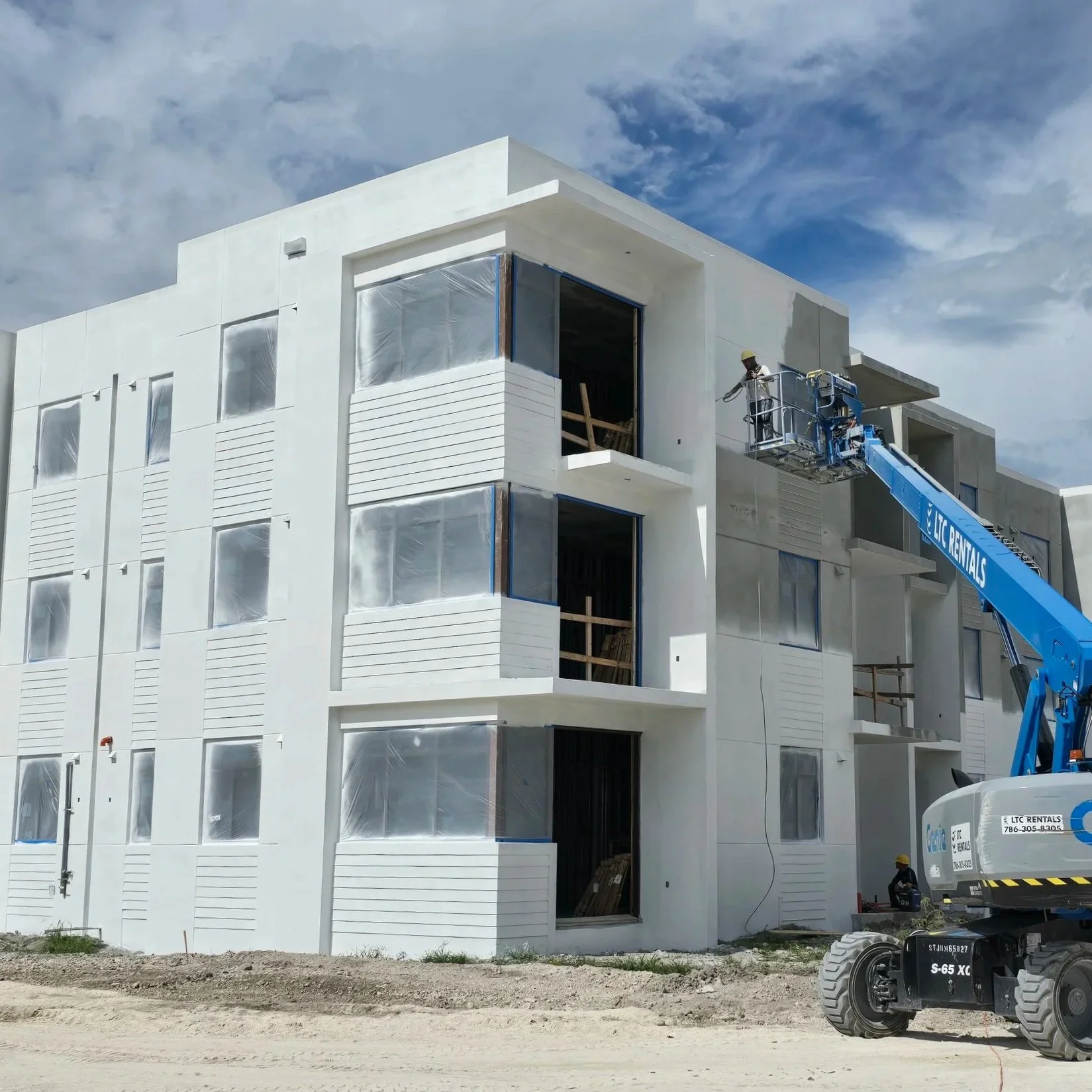 Construction workers using a blue lift to paint or work on the exterior of a modern white apartment building with multiple balconies, some covered with protective plastic, under a partly cloudy sky.