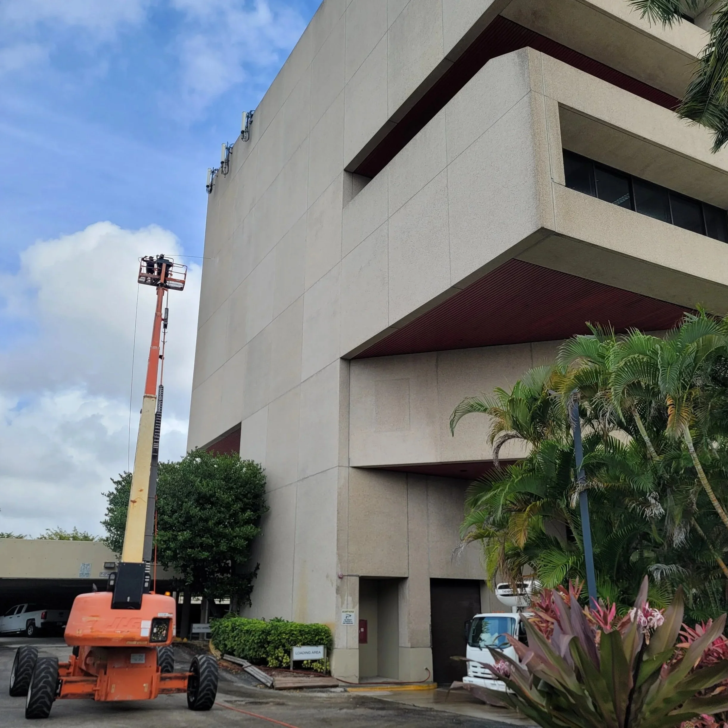 A multi-story beige building with brown accents, a large tree, palm trees, and colorful plants in the foreground. There is a lift truck and a white vehicle parked near the building.