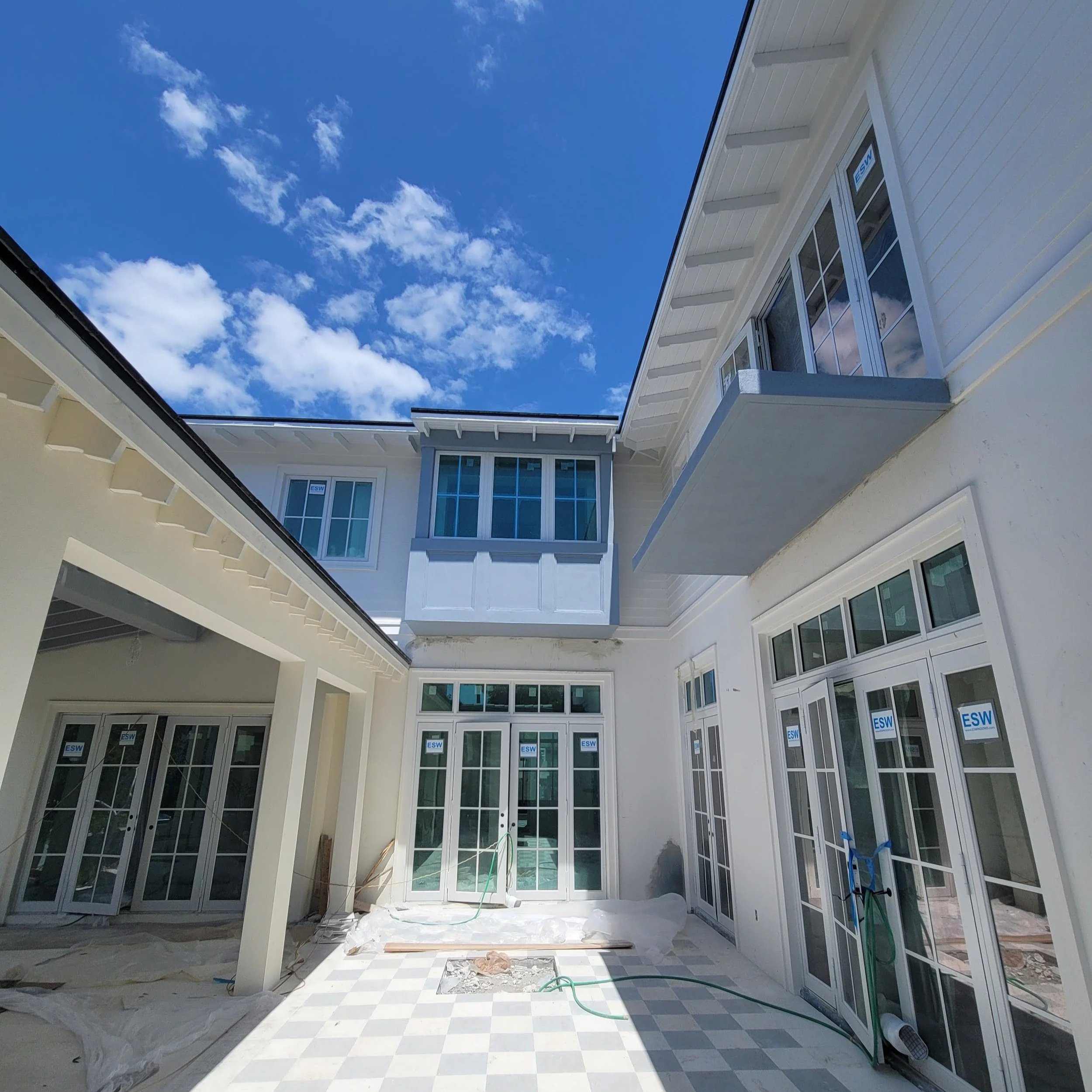 View of a modern Miami house, with white paint, under construction and being painted with multiple large windows and a bright blue sky with white clouds.