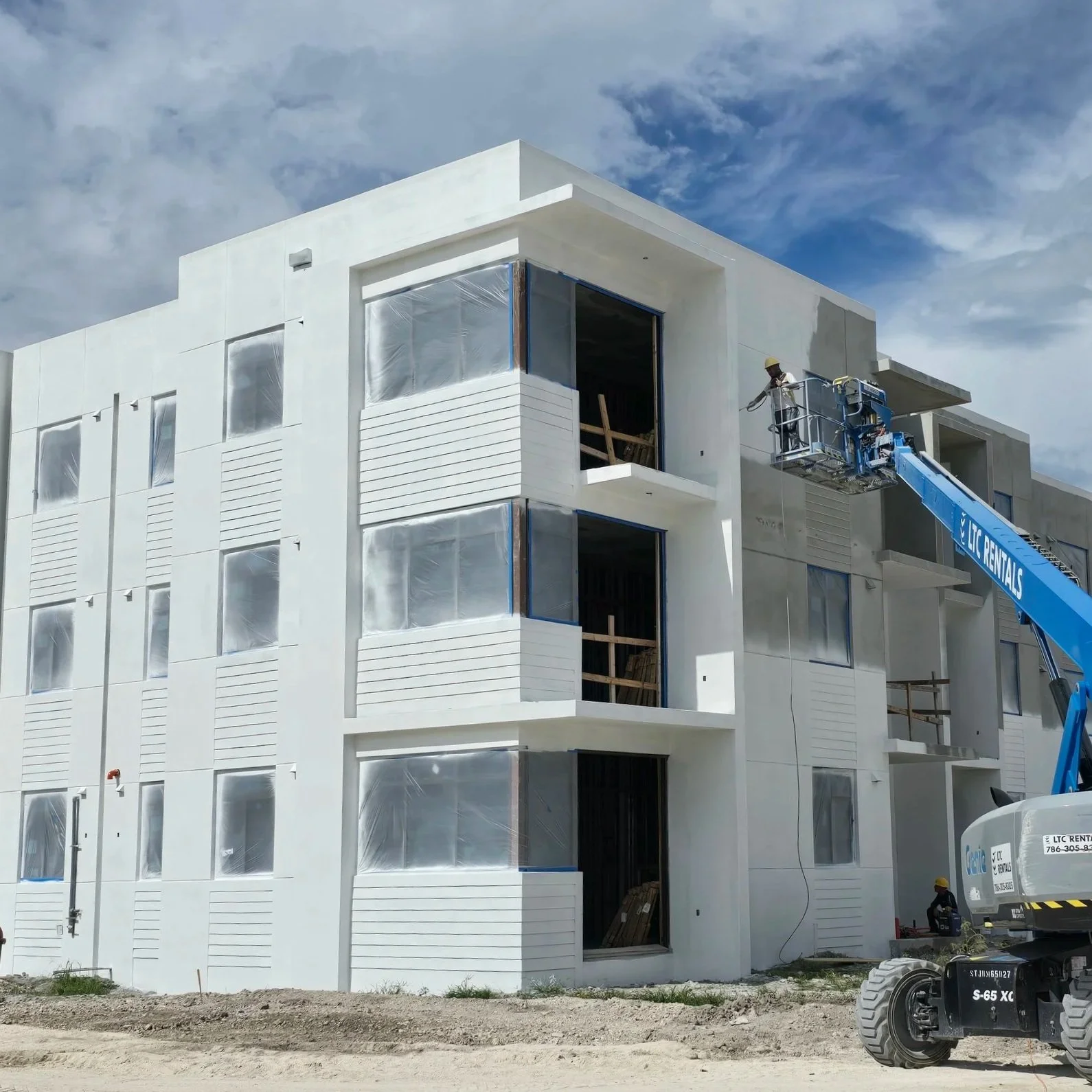 Expert paint workers painting a white multi-story residential building in Miami with balconies, using a crane lift, under partly cloudy sky.