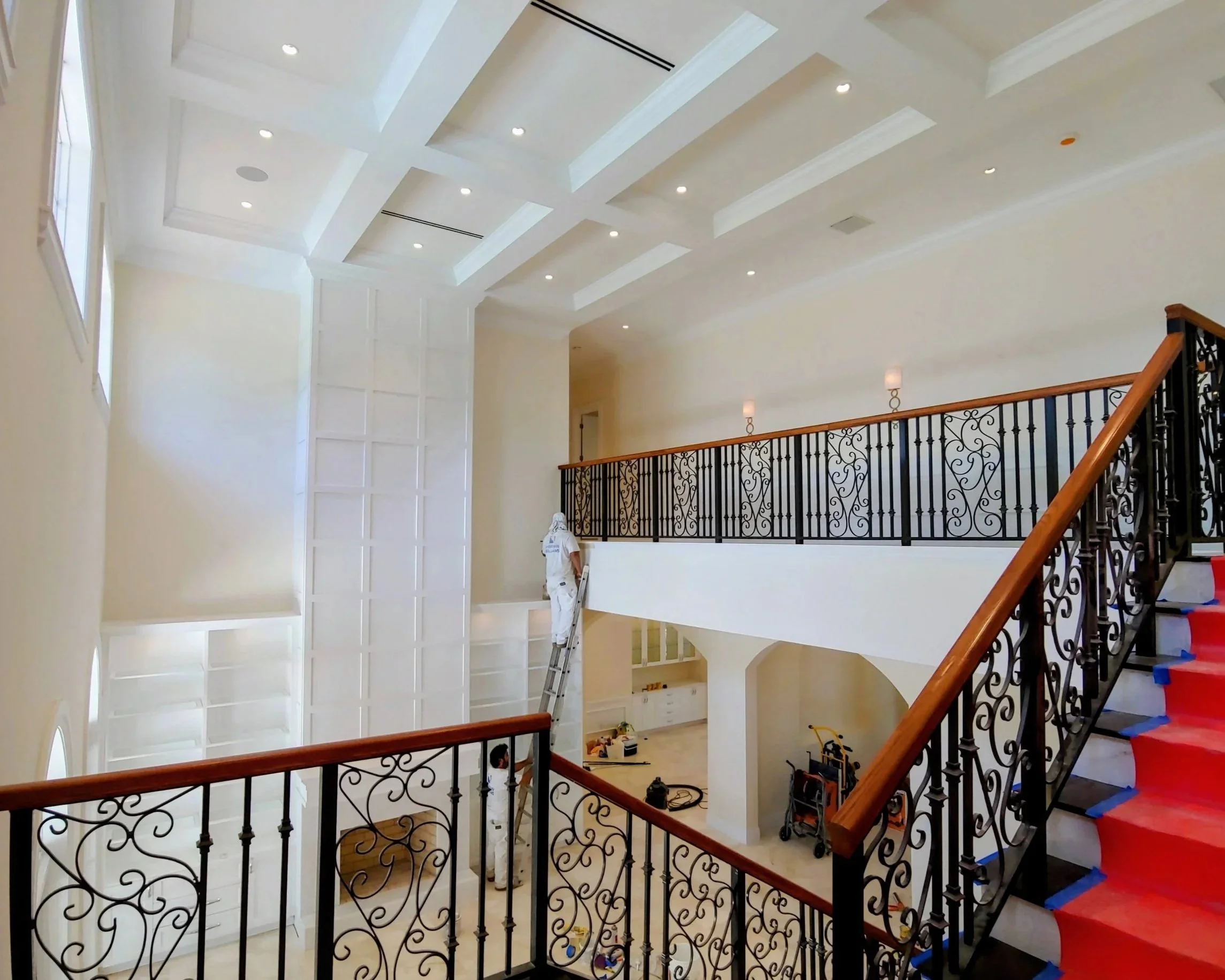 Interior of a spacious, two-story house with white walls, coffered ceiling, and decorative wrought iron balcony railings. A worker in white attire is standing on a ladder, interior is under construction or renovation.