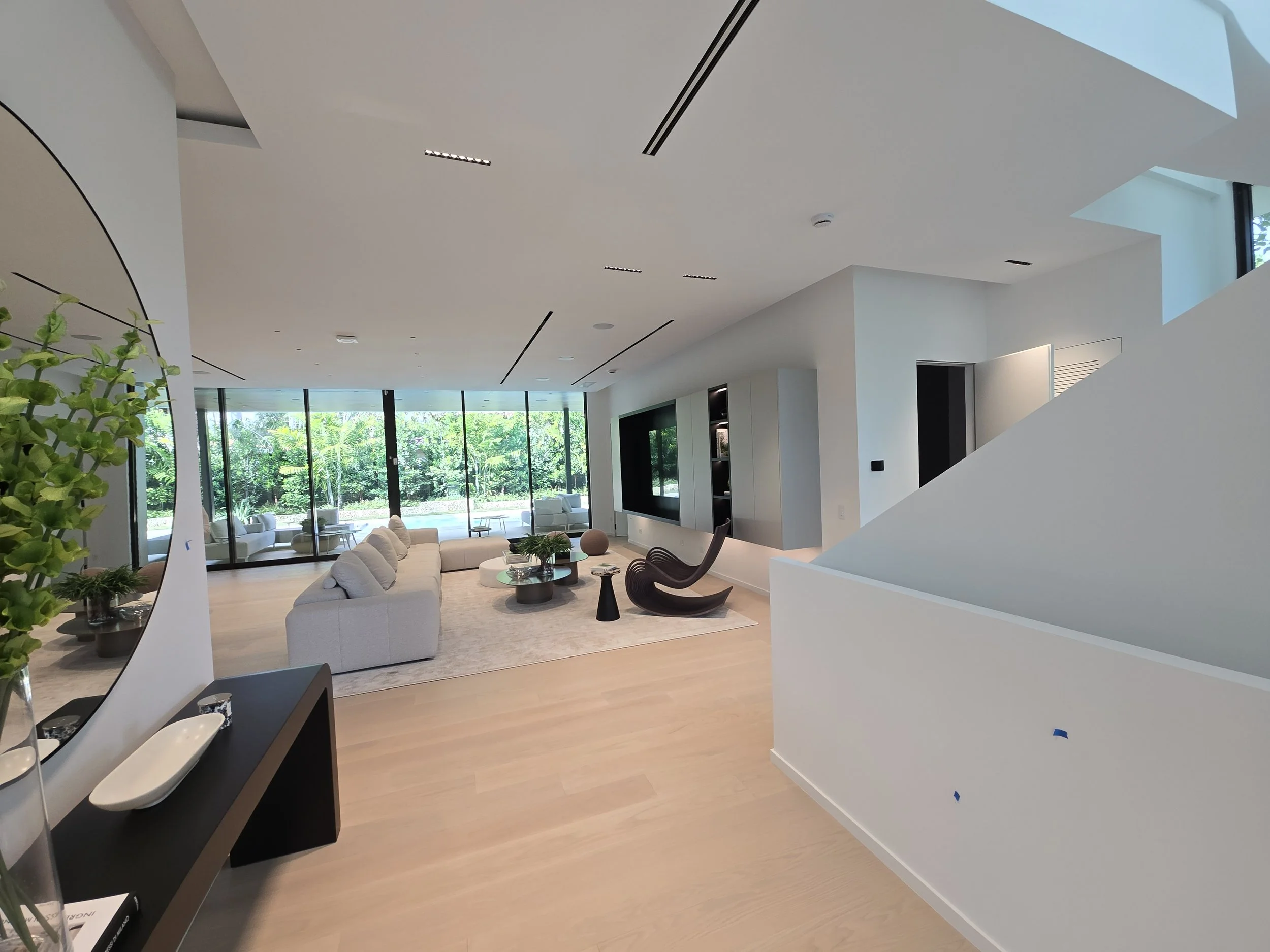 Modern living room with large glass windows, white sofa, black lounge chair, round coffee tables, and green plants, filled with natural light.