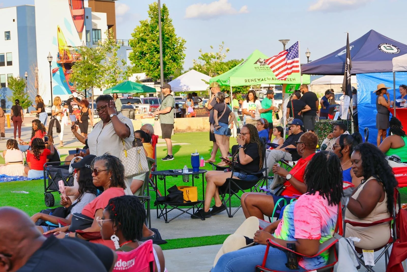 A diverse group of people gathered outdoors at a community event, sitting in chairs and standing near tents on a grassy area with city buildings and trees in the background.