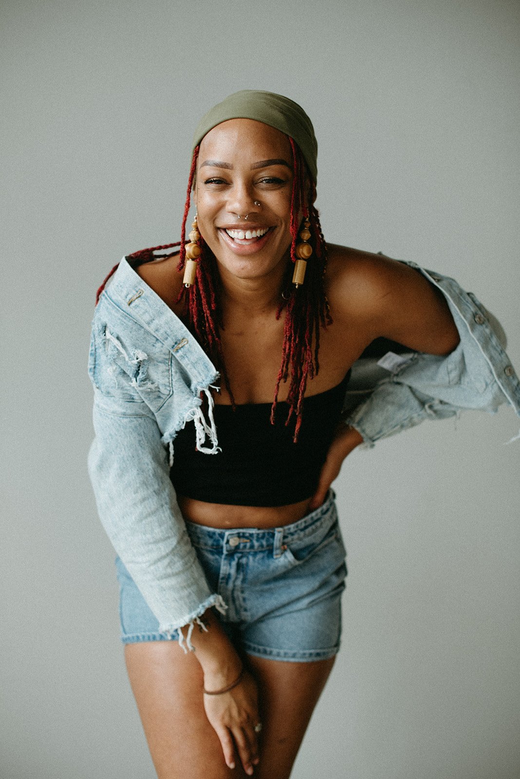 Young woman with red dreadlocks wearing a green headscarf, earrings, a black crop top, and a light denim jacket and shorts, smiling and posing with one hand on her hip.