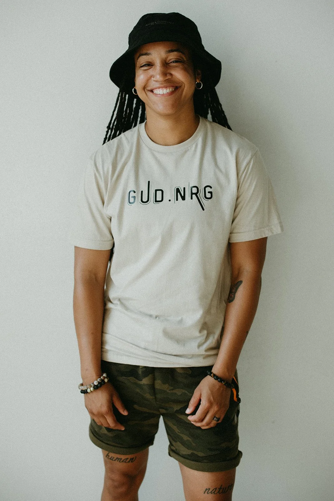 A woman smiling, wearing a black bucket hat, earrings, a beige T-shirt with black lettering, camo shorts, bracelets, and a ring, standing against a plain light-colored wall.