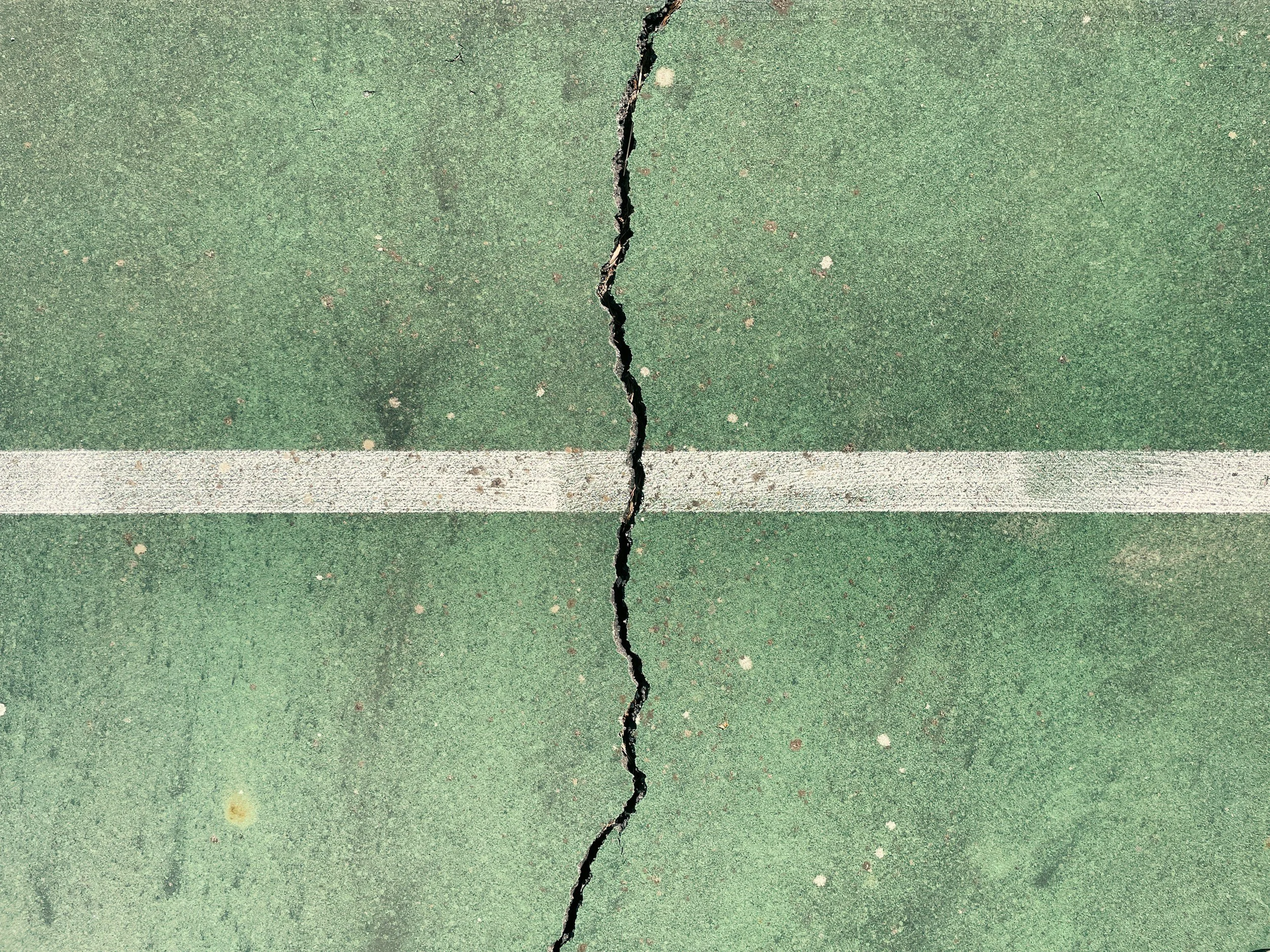 A green outdoor sports court surface with a large vertical crack running through the middle, and a white boundary line near the bottom.