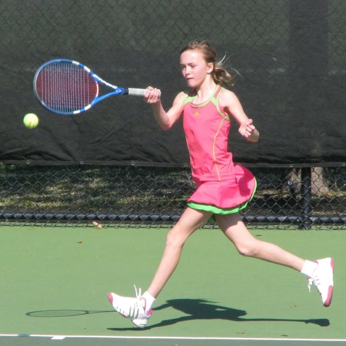 Young girl in a pink sports dress playing tennis on a green court.
