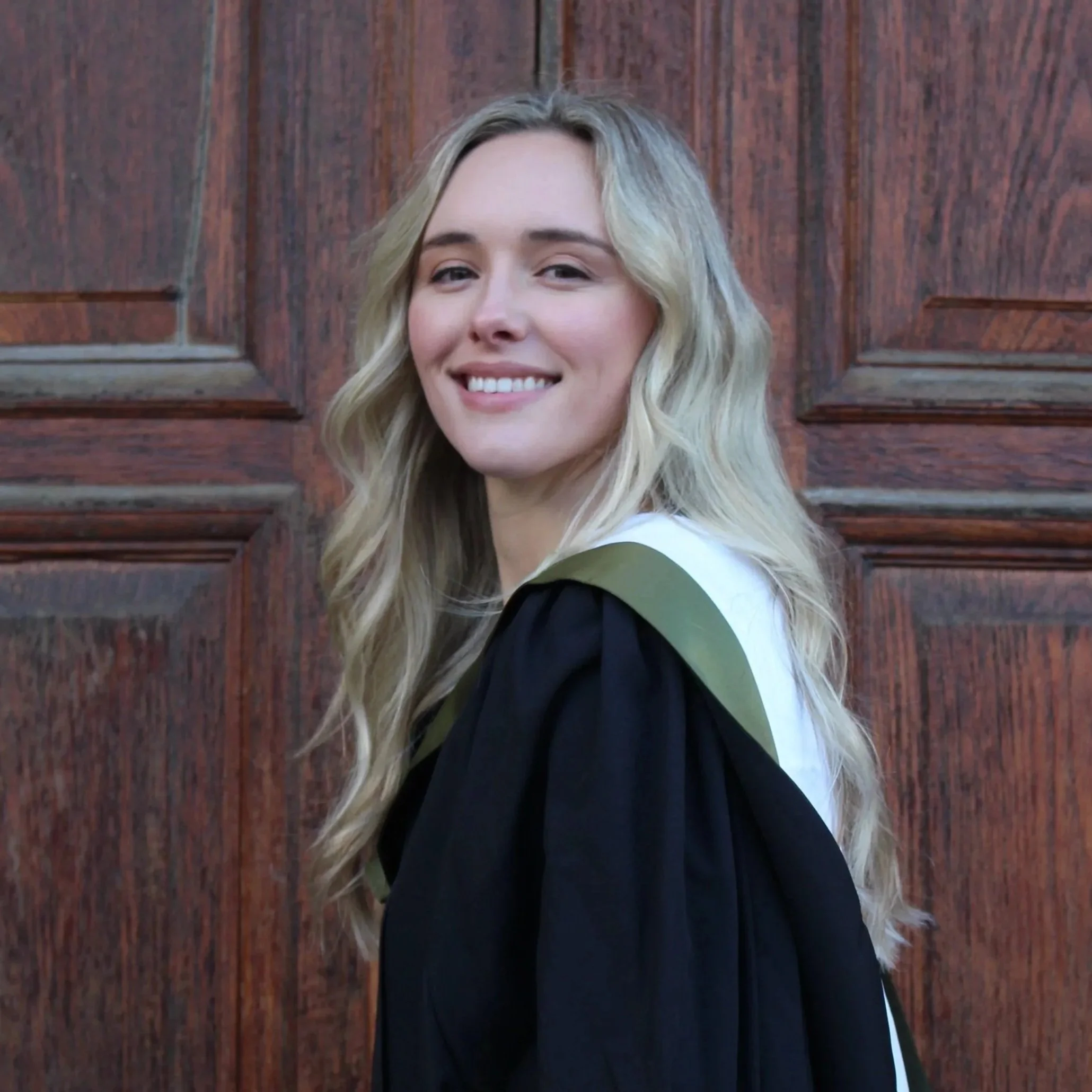 A young woman with long, wavy blonde hair smiling, standing outdoors in front of a wooden wall.