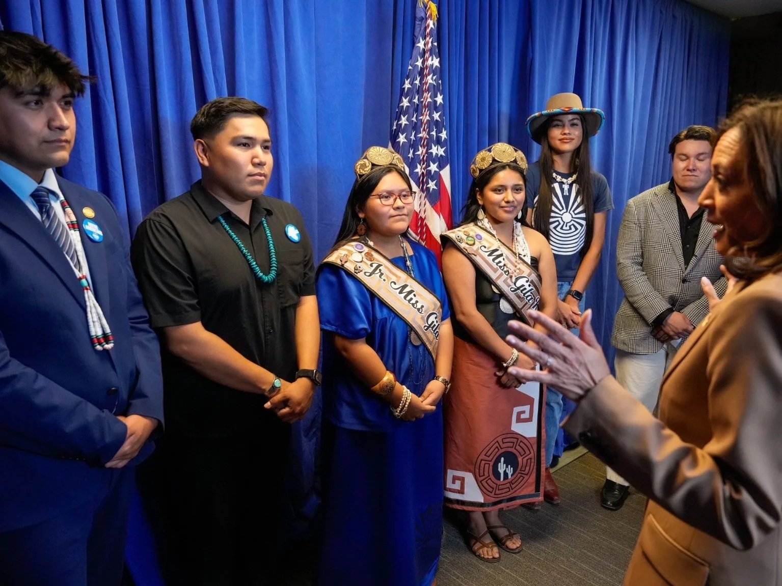 Group of young women and men standing in a line during a ceremony, with some wearing sashes reading "Miss Gila" and "Jr. Miss Gila," colorful attire, some with beaded jewelry and traditional accessories, standing in front of a blue curtain and American flag, as a woman with brown hair speaks to them.