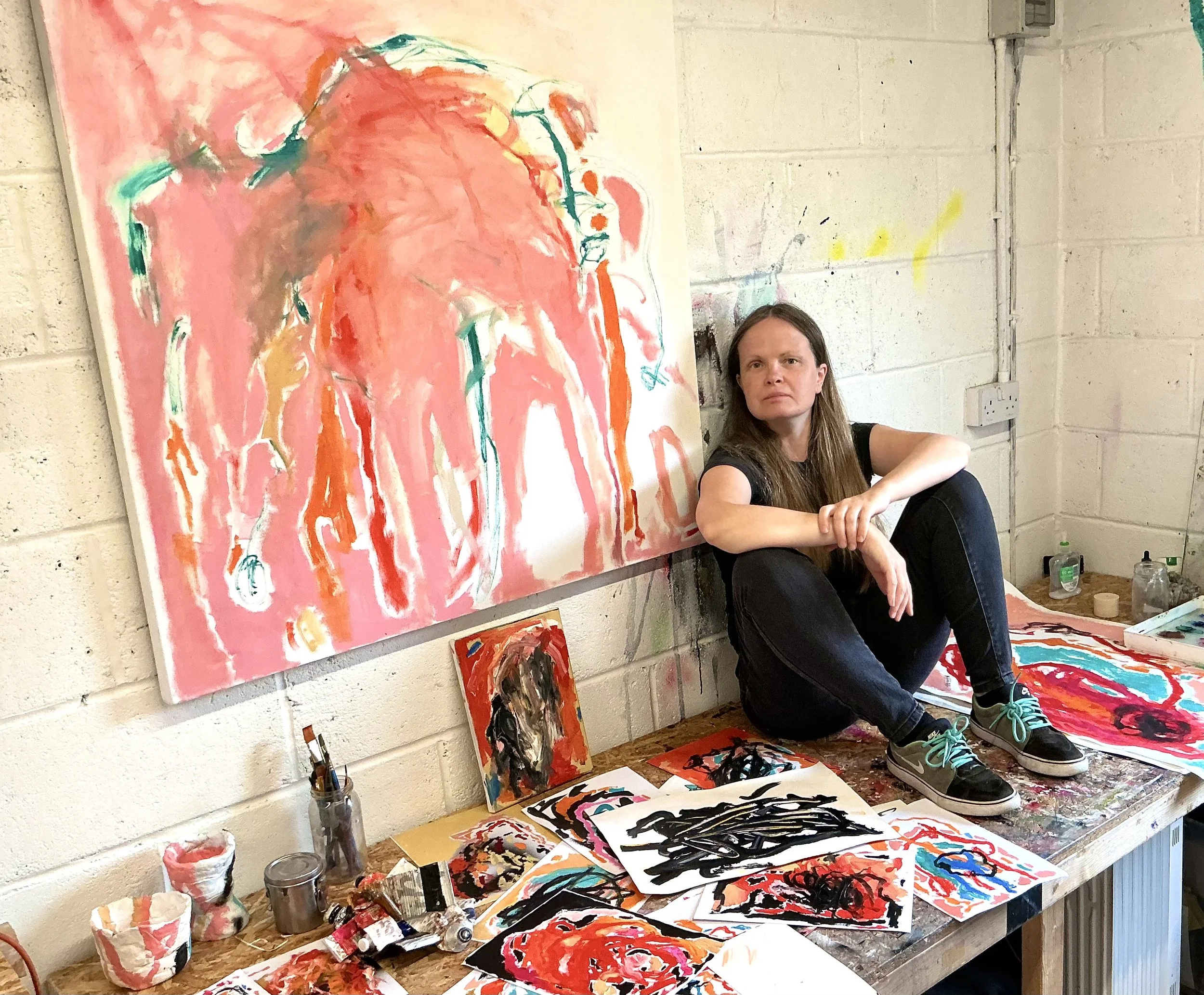 A woman sitting on the floor next to a large abstract painting with bright pink, orange, and teal colors. The painting is on a white brick wall, with art supplies and smaller paintings on the table below.