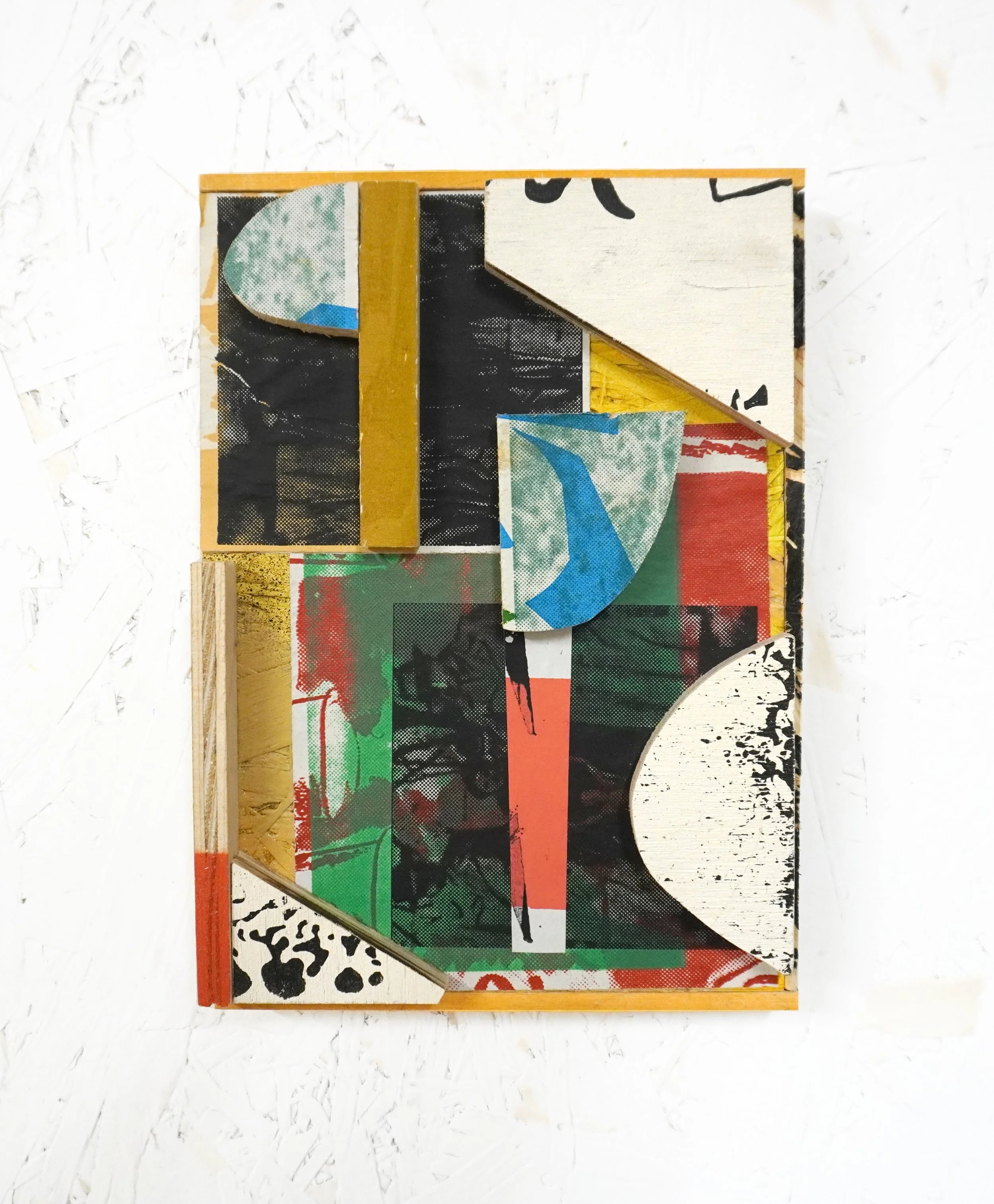 An abstract mixed media art piece featuring layers of paper, paint, and collage elements in black, white, red, green, blue, and yellow, mounted in a wooden frame, displayed on a white textured surface.