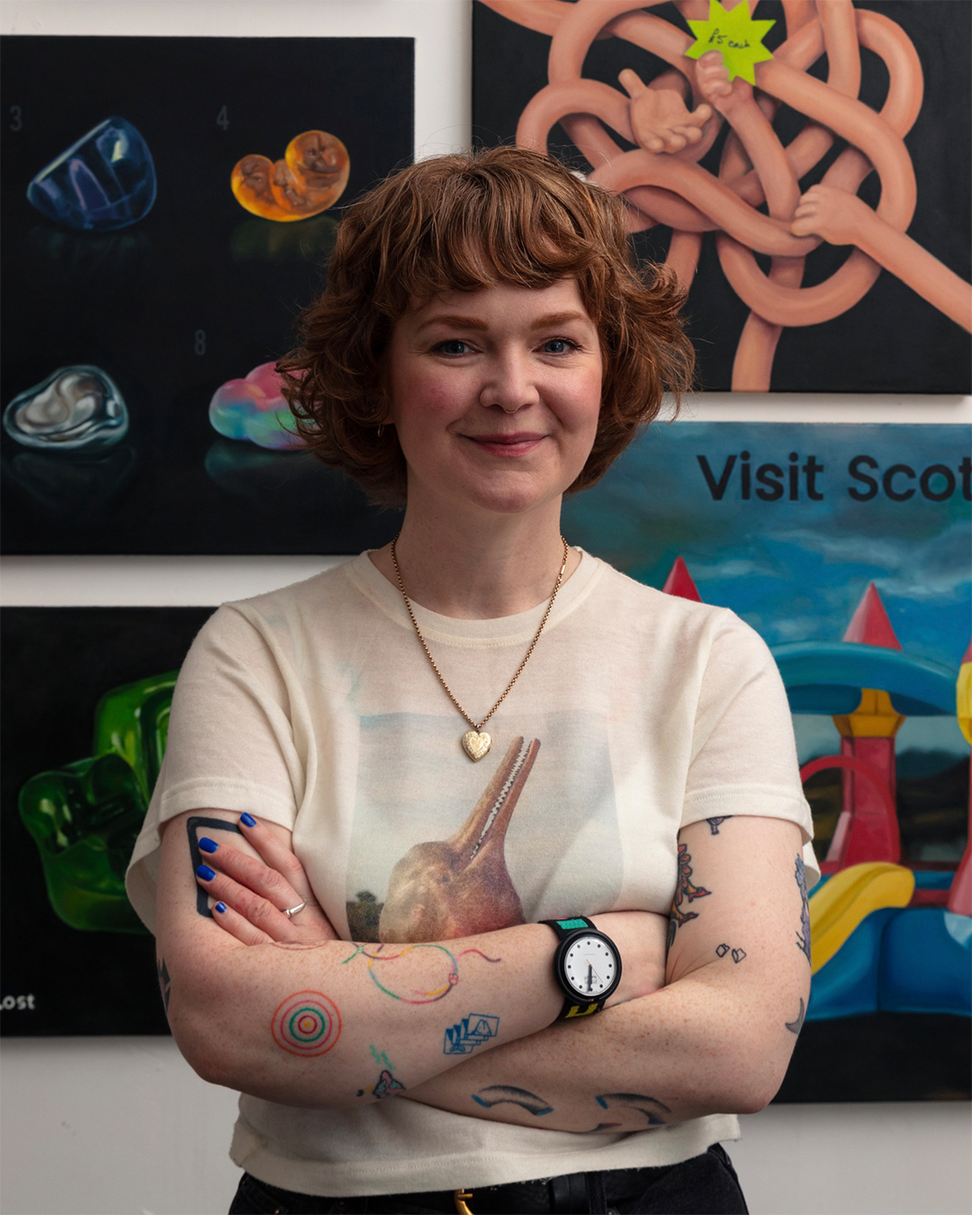 A woman with short curly red hair smiling and crossing her arms, standing in front of colorful artwork including a sign that says "Visit Scot" and paintings of abstract objects and a knot with arms.