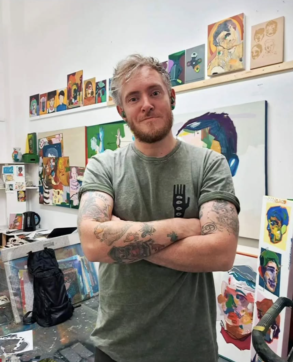 A man with arms crossed and tattoos standing in an art studio filled with colorful paintings and artwork on the walls and table.