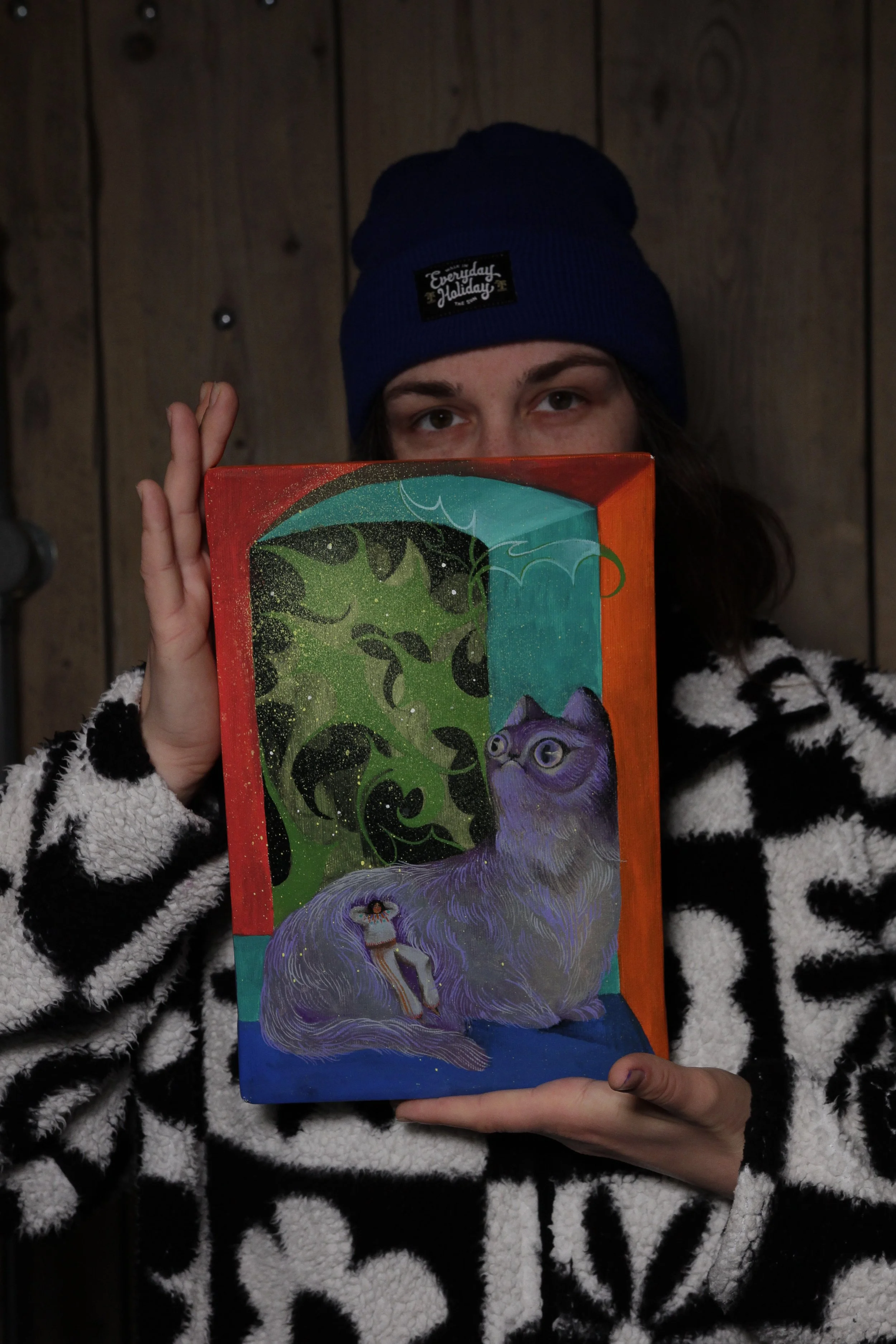 A person wearing a navy blue beanie with a small patch that reads "Everyday Holiday" and a black and white patterned fleece jacket, holding a colorful illustrated book in front of their face against a wooden wall background.