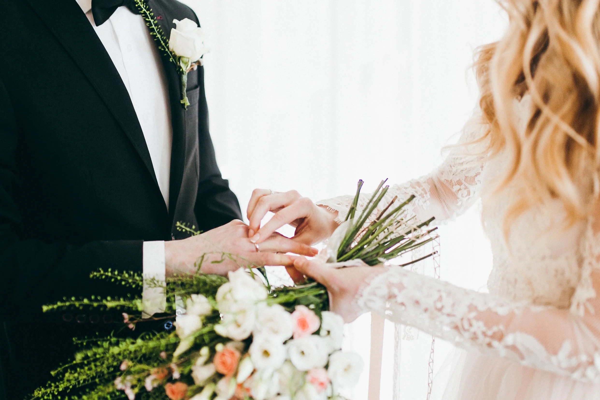 A bride and groom exchanging rings during a wedding ceremony, with the bride wearing a lace dress and the groom in a black tuxedo with a white shirt and bow tie, surrounded by floral arrangements.