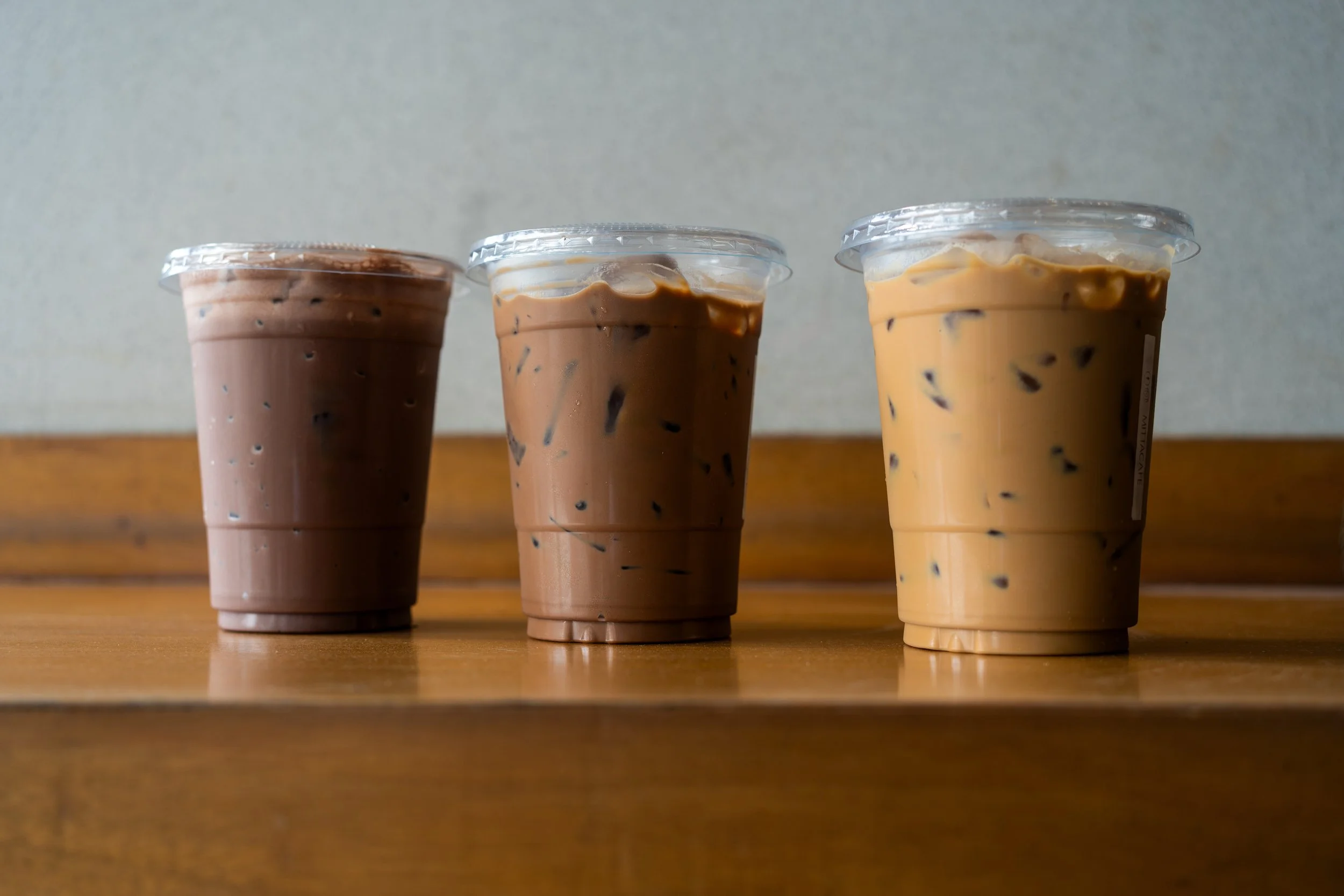 Three plastic cups filled with iced coffee of different shades, placed on a wooden surface. The cups have clear lids and visible ice cubes inside.