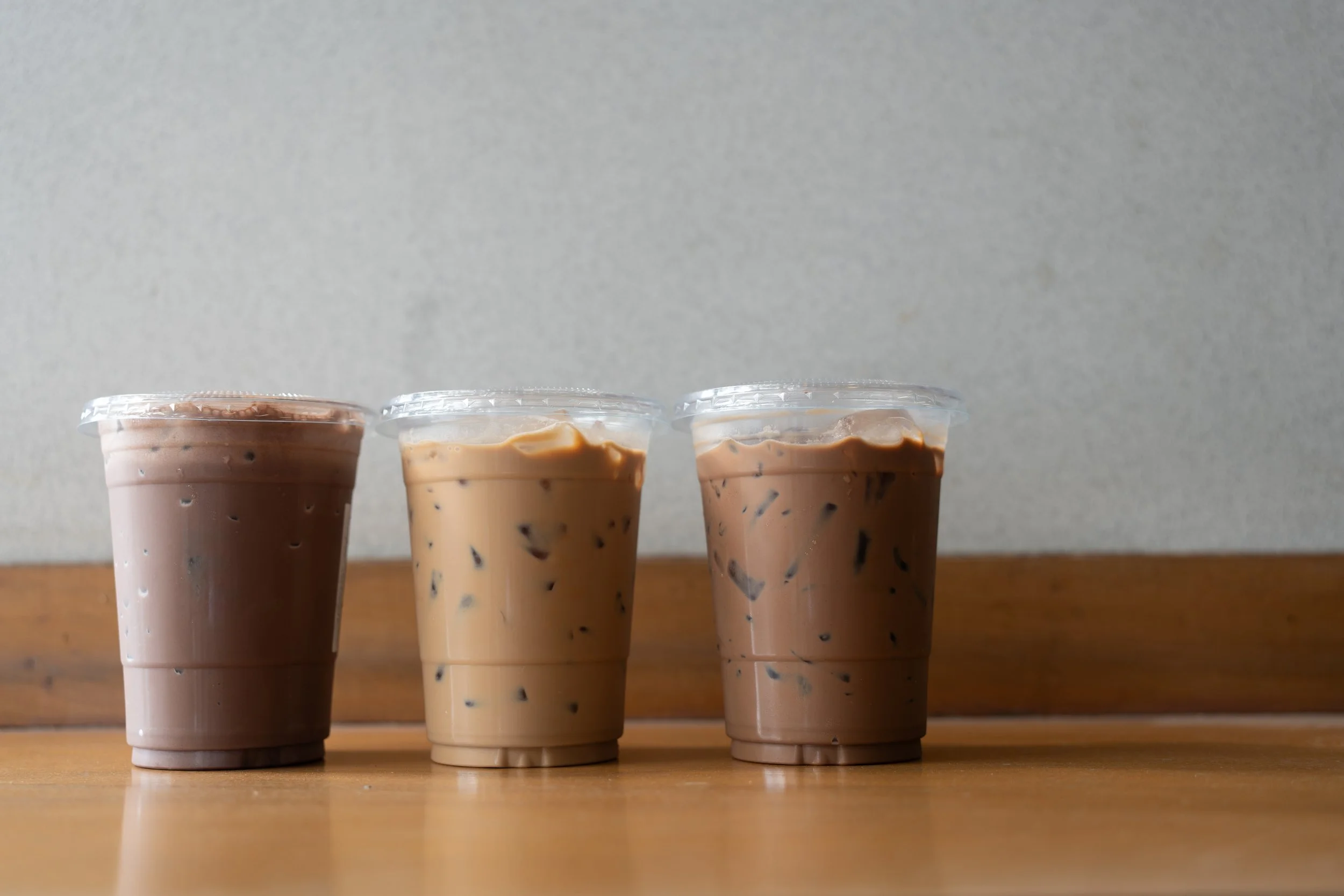 Three transparent plastic cups filled with iced coffee beverages arranged on a wooden surface in front of a plain gray wall.