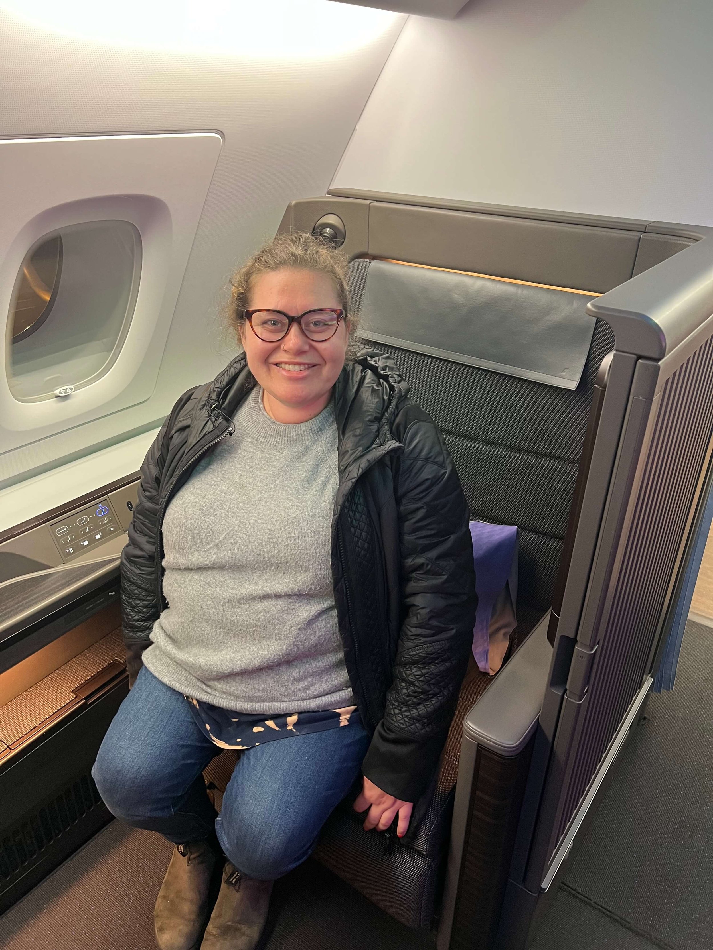 Sophie Keller is sitting in an airplane seat next to a window, smiling at the camera, wearing glasses, a gray sweater, a black jacket, jeans, and boots.