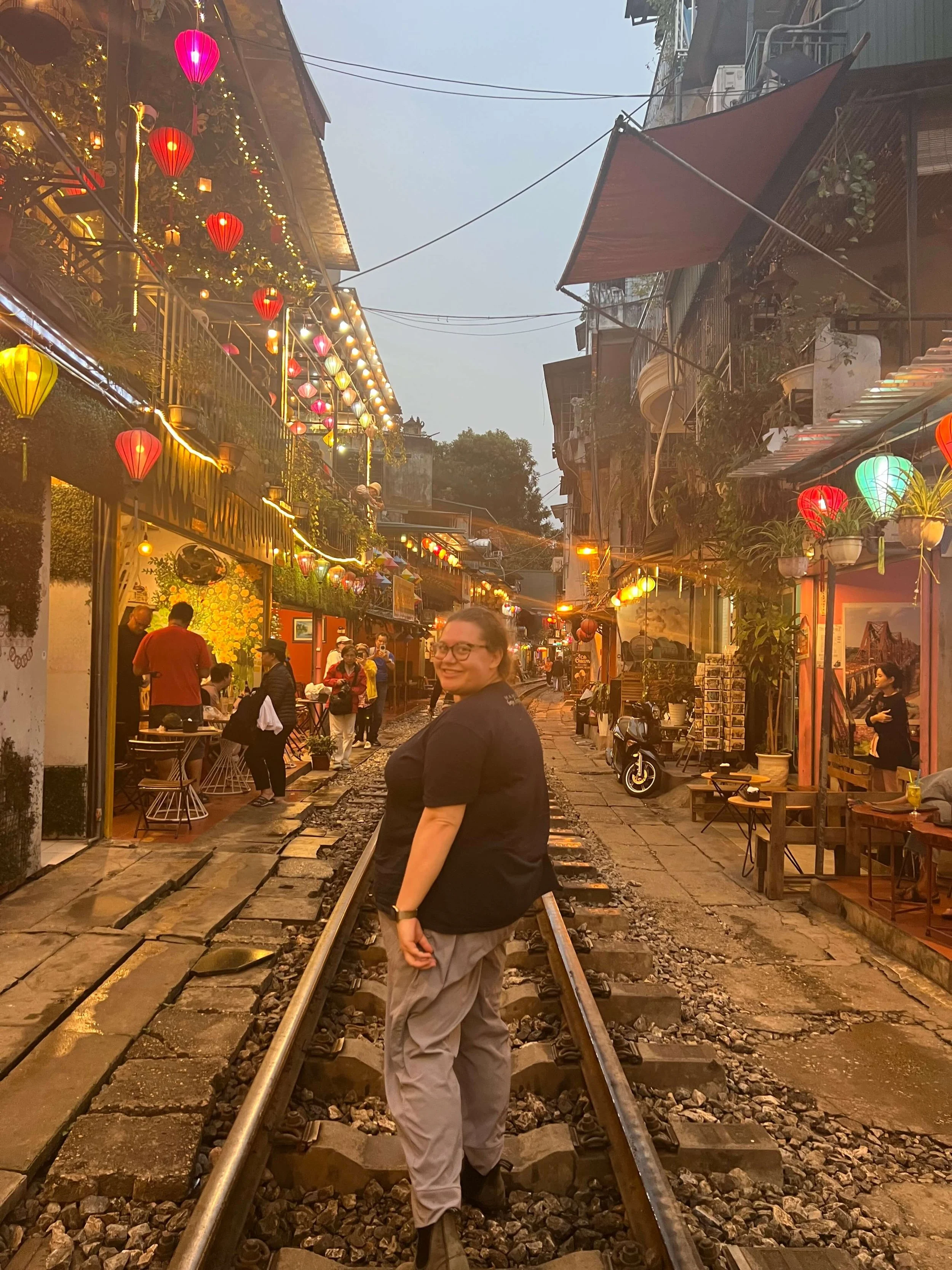 Sophie Keller is smiling while standing on railway tracks inside a lively, colorful street with hanging lanterns, small shops, and outdoor seating in Vietnam