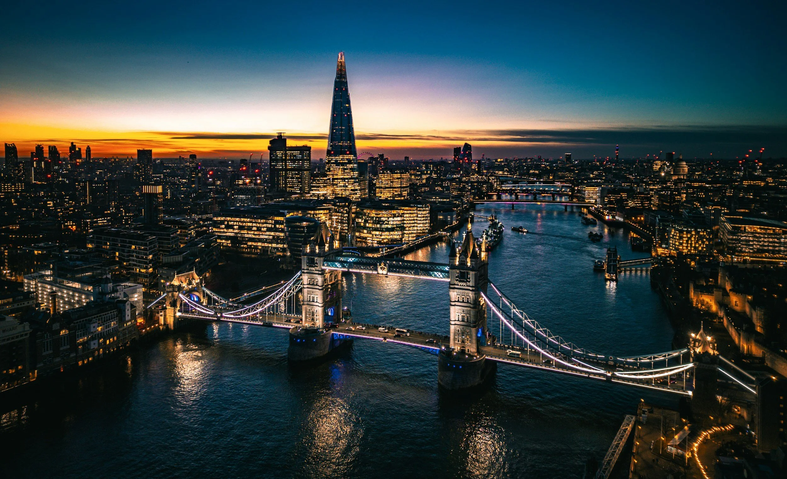 Aerial view of London at sunset featuring Tower Bridge illuminated over the River Thames with the Shard skyscraper in the background and city lights.