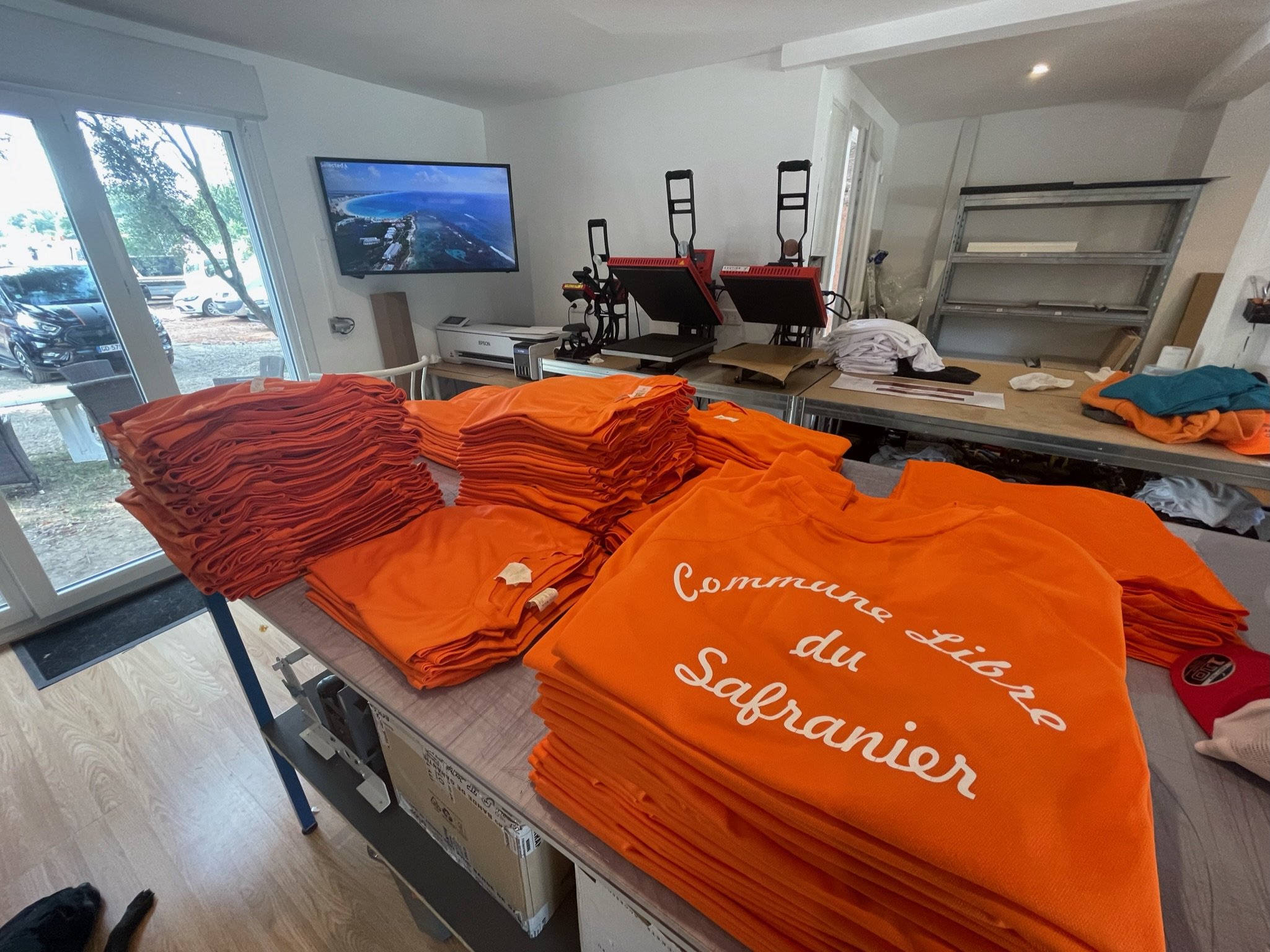 Table avec de nombreuses t-shirts orange pliés, un mur avec une poignée de camping, une télévision murale, des fenêtres laissant entrer la lumière, et un mur avec des étagères métalliques contenant divers objets. Envahissement de matériaux et équipement, probablement une salle de préparation ou de stockage pour un événement.