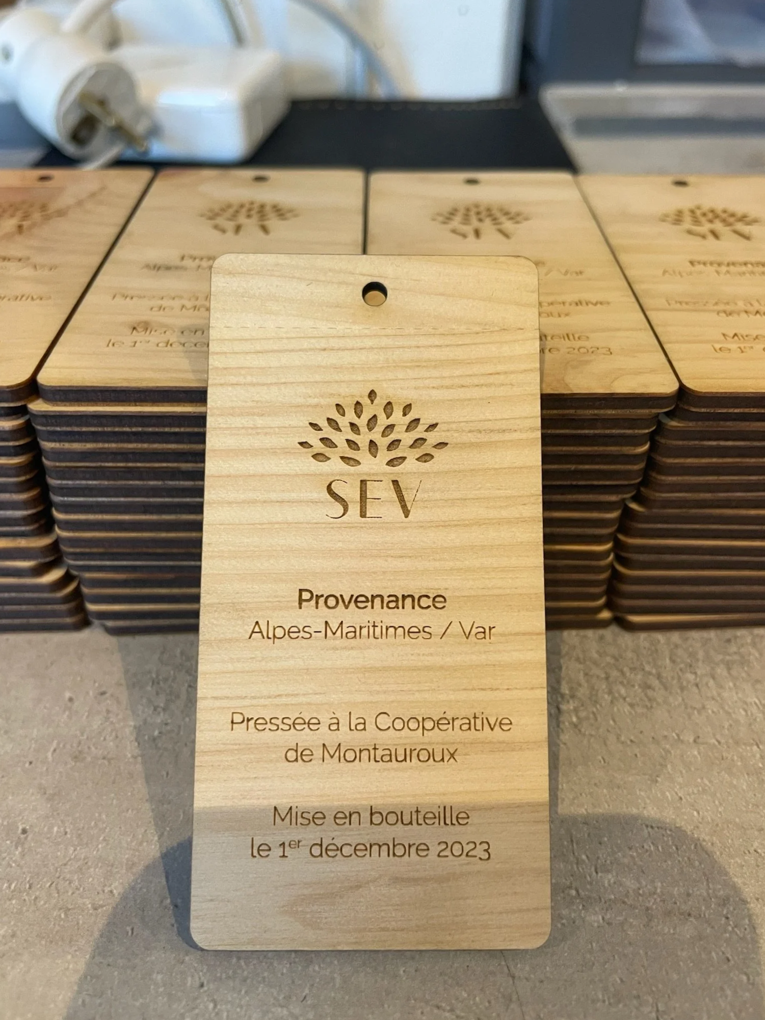 Tags en bois gravés avec le logo d'une arbre et le texte : SEV, Provenance Alpes-Maritimes / Var, Pressée à la Coopérative de Montauroux, Mise en bouteille le 1er décembre 2023.