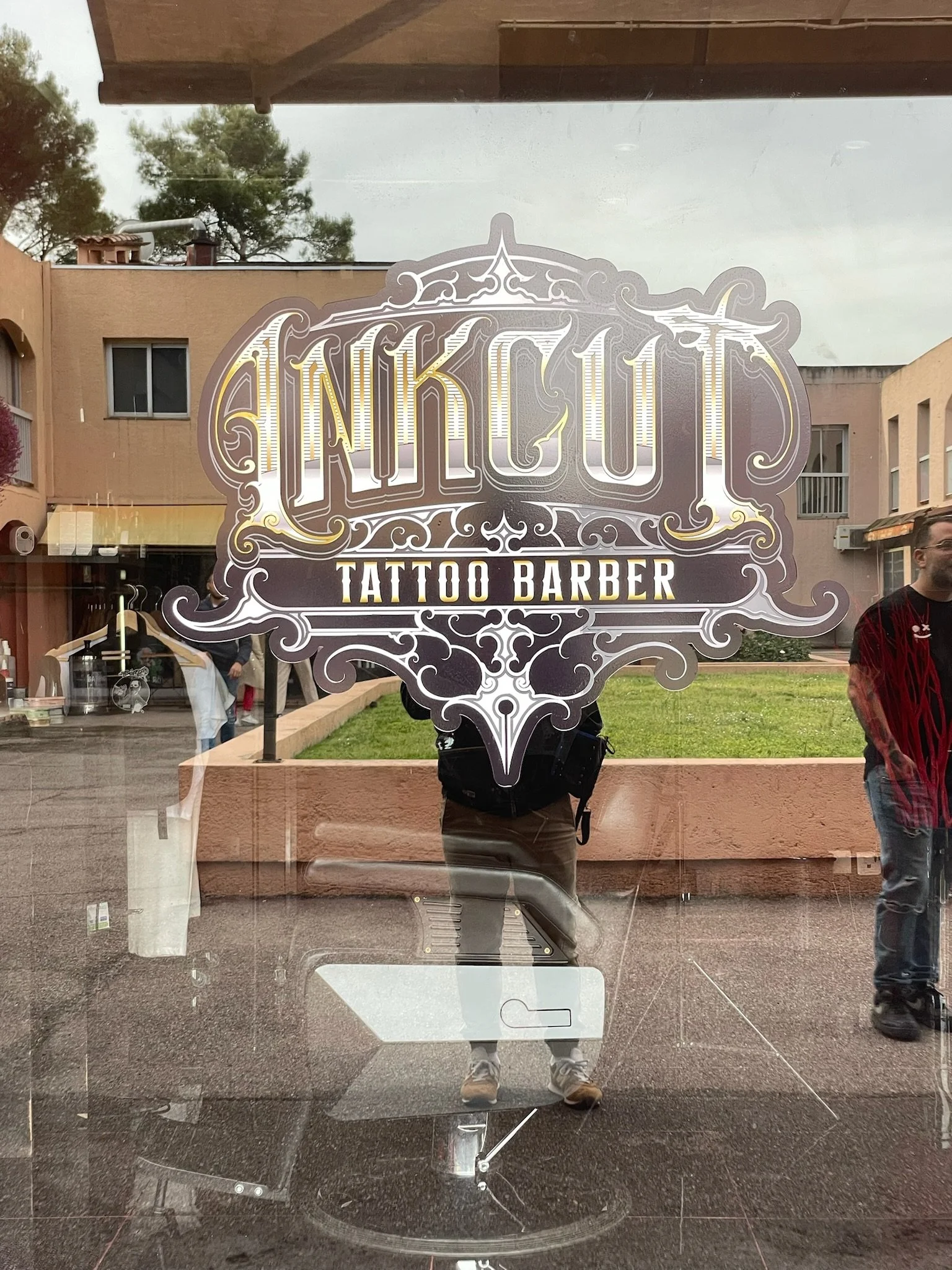 Une porte en verre avec un logo de salon de tatouage et barber shop, reflet de la rue avec des personnes et des bâtiments.