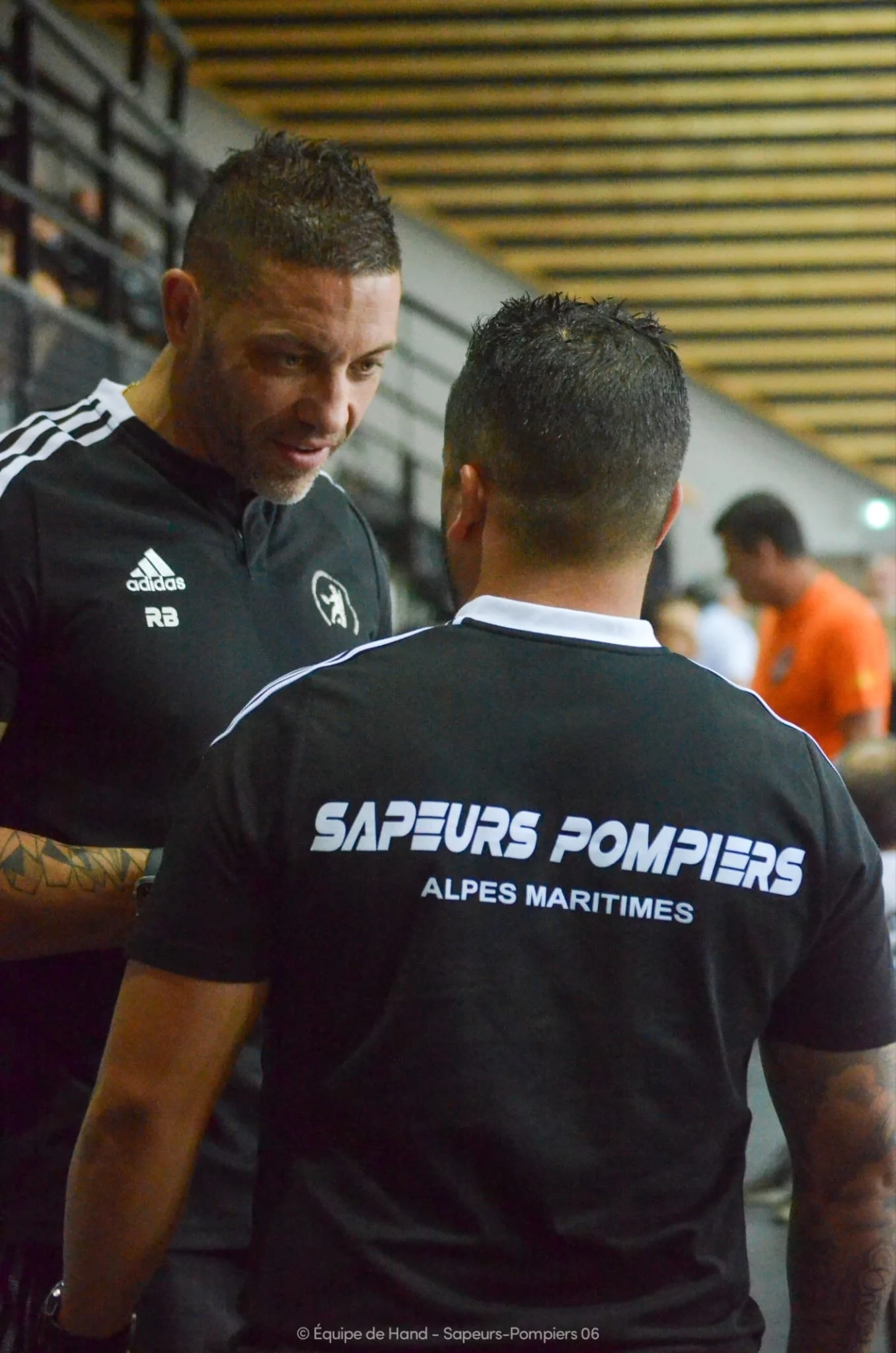Deux personnes en uniforme de pompiers discutent, avec un arrière-plan d'une salle intérieure. Le dos d'une personne porte un t-shirt avec l'inscription 'SAPEURS POMPIERS ALPES MARITIMES'.