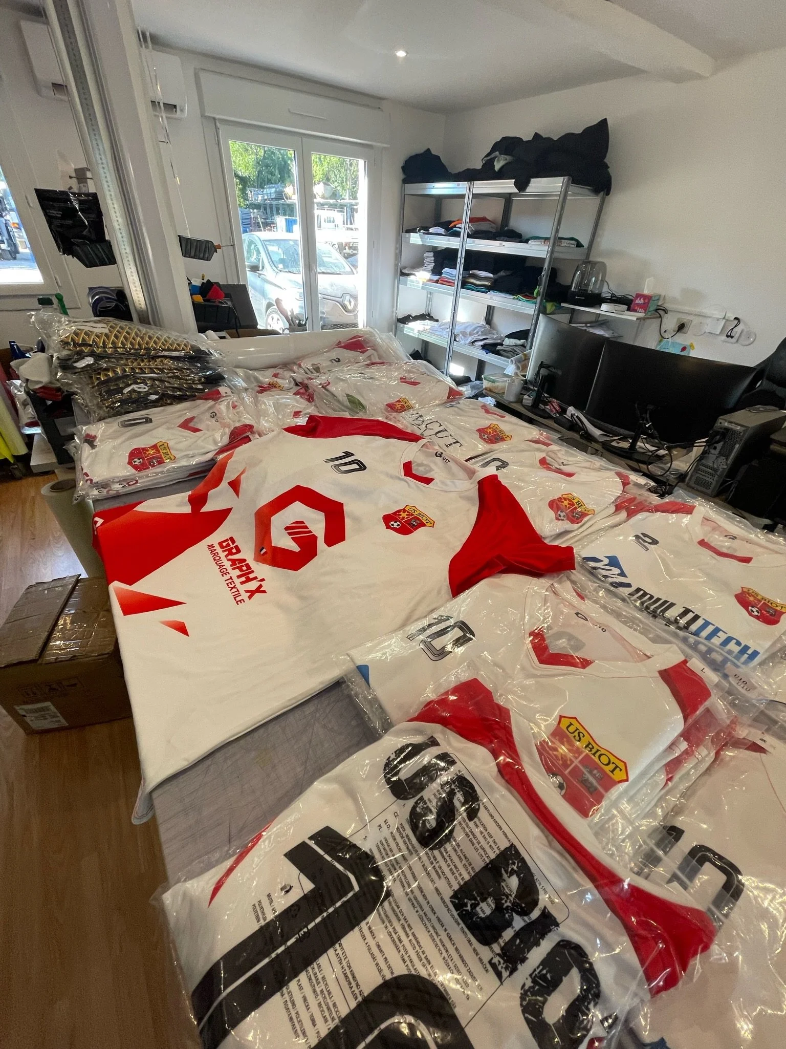 Une table recouverte de plusieurs paquets plastifiés de maillots de football, probablement de l'équipe US BIOUS, dans une pièce avec étagère en métal, un ordinateur et une porte fenêtre donnant sur l'extérieur.